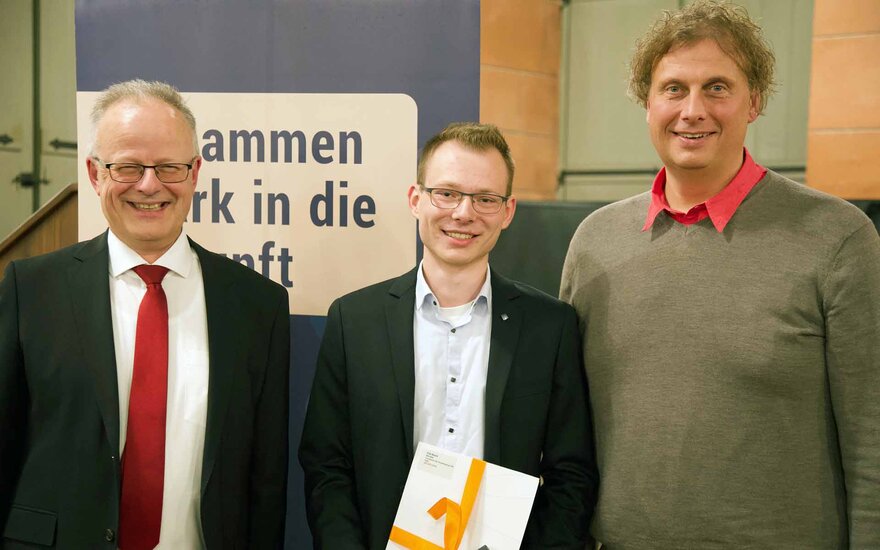 Drei Männer stehen lächelnd nebeneinander. Der junge Mann im schwarzen Sacko in der Mitte hält einen Umschlag mit einer gelben Schleife in der Hand.