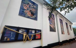 Vier große Wandgemälde mit den Badenden in den Farben Blau, Türkis und Rosa hängen an der weiße Außenfassade des Filmtheaters. Darunter ein breiter Schaukasten mit einer goldenen Schleife darum, in welchem Plakate über das Leben und Wirken des Künstlers hängen