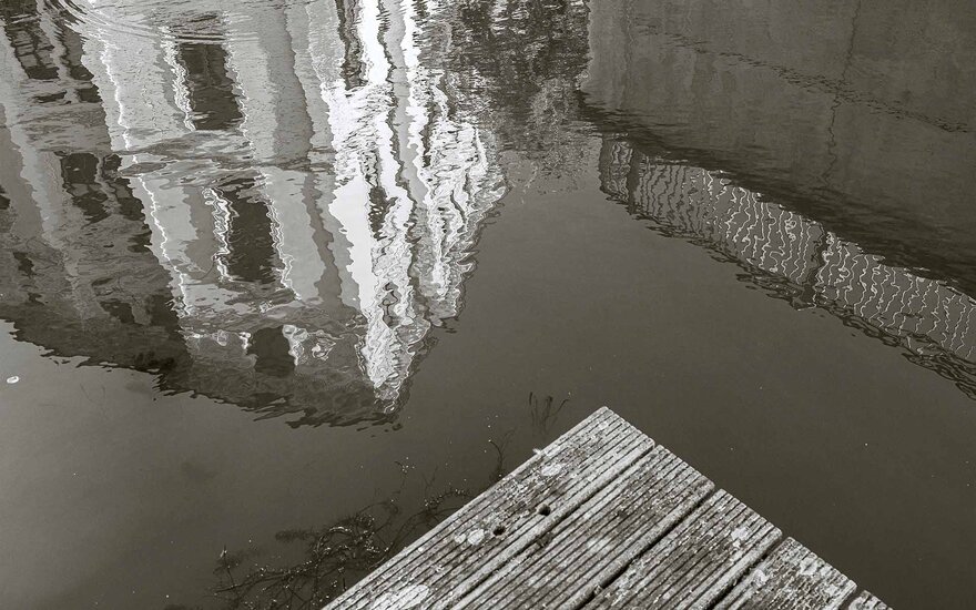 dreieckiger Teil eines Holzplateaus im Wasser, in dem sich ein Gebäude und eine Wand spiegeln