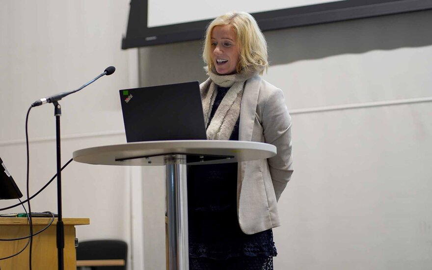 Eine blonde Frau steht an einem Rednerpult hinter einem Laptop und spricht ins Mikrofon.