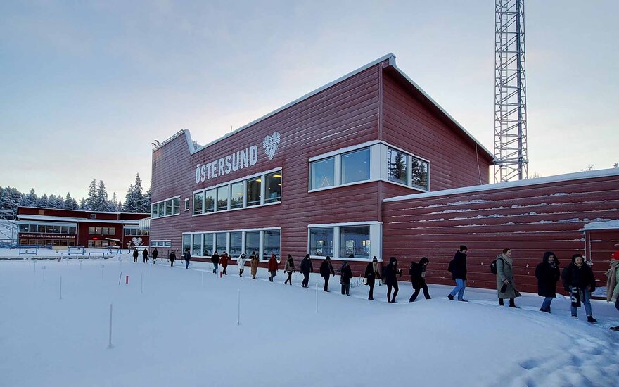 Gebäude aus braunem Holz mit Flachdach, vor dem eine Reihe von Personen hintereinander durch den Schnee läuft