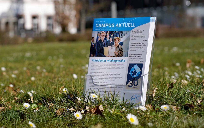 Hochschulmagazine im Aufstelle auf Wiese mit Gänseblümchen vor Gebäuden der TH