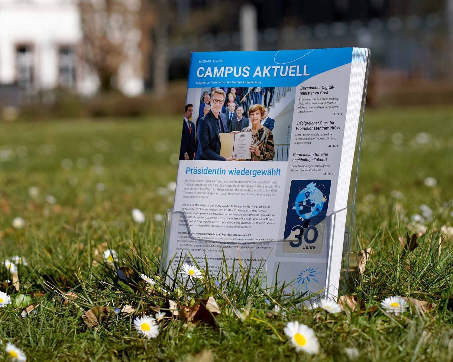 Hochschulmagazine im Aufstelle auf Wiese mit Gänseblümchen vor Gebäuden der TH
