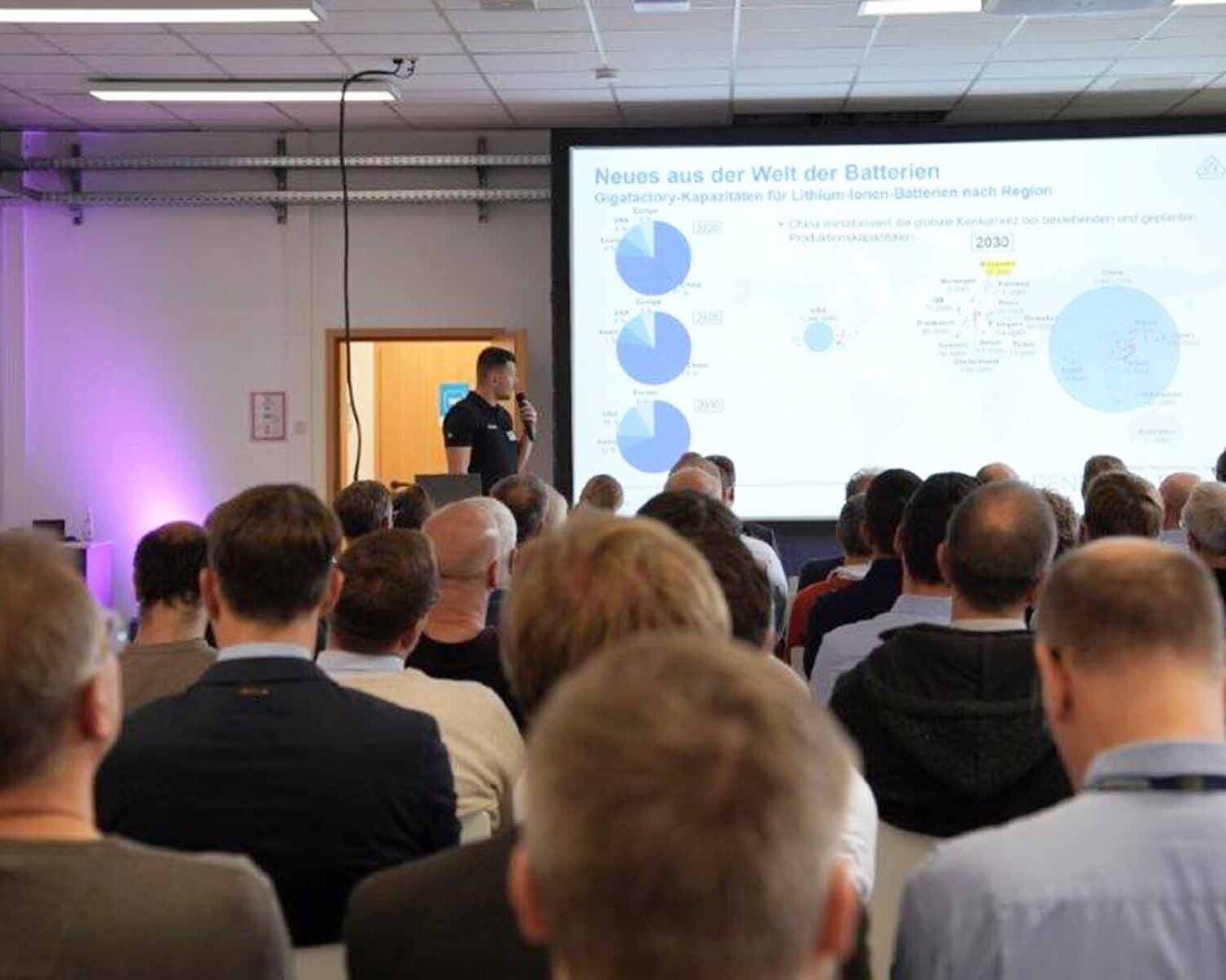 In Reihen sitzende Menschen von hinten mit Blick auf einen Referenten und eine Präsentation.