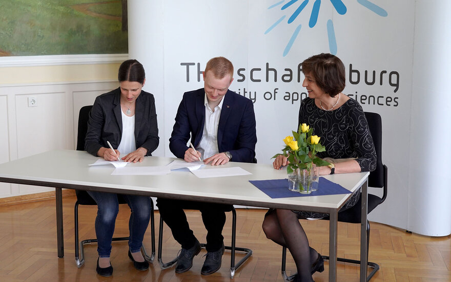 Drei Personen sitzen an einem Tisch vor einer Messewand mit TH-Aschaffenburg-Logo und unterzeichnen einen Vertrag