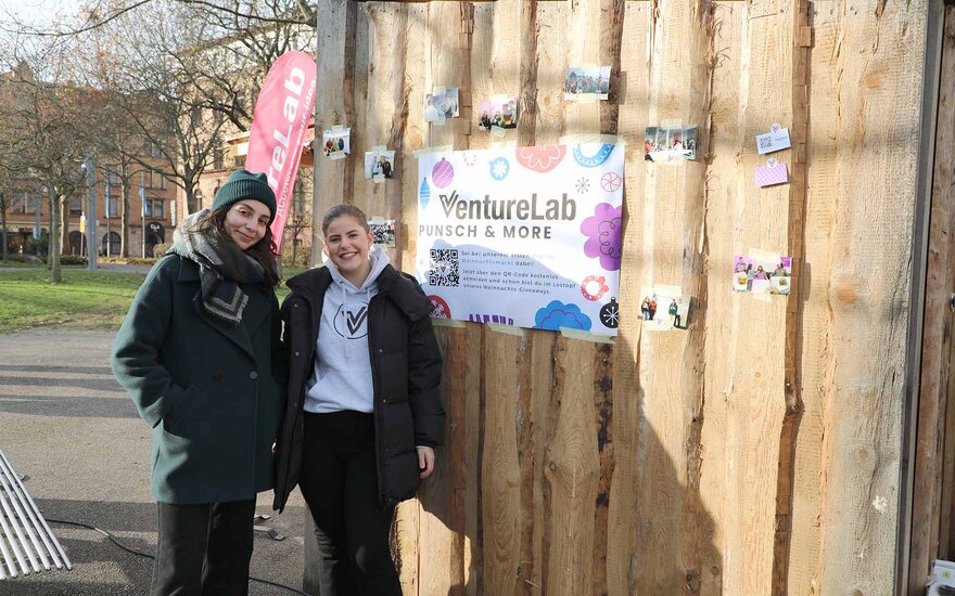 Zwei junge Frauen stehen vor einem Holzstand mit einem Venture-Lab-Plakat und lächeln in die Kamera