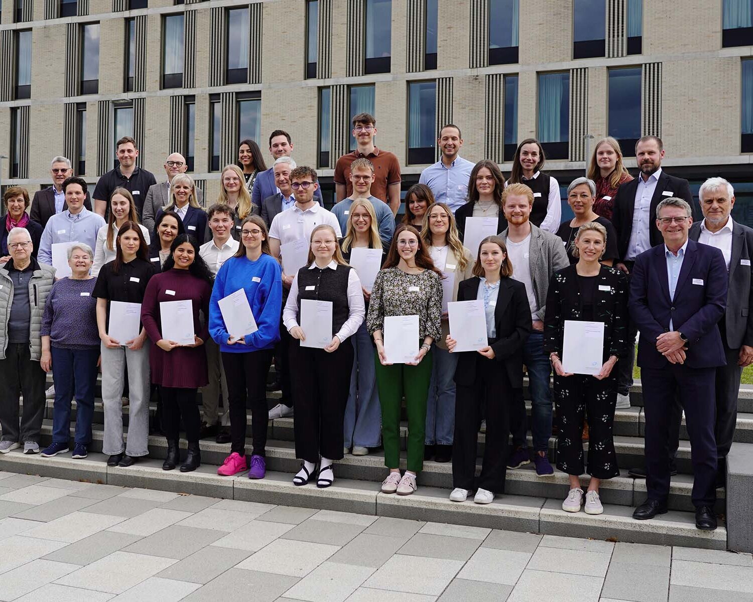 Gruppenbild mit rund 40 Personen, die teilweise Zertifikate halten, auf Treppenstufen vor den Neubauten