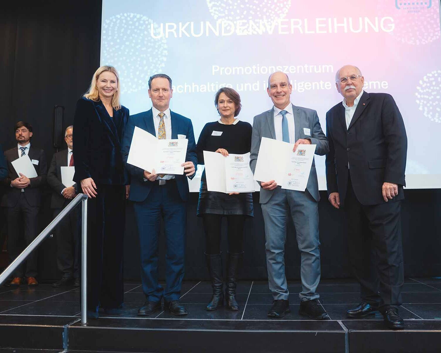 Fünf Menschen stehen vor einer Leinwand mit dem Schriftzug Urkundenverleihung. Drei Personen halten Urkunden in den Händen.