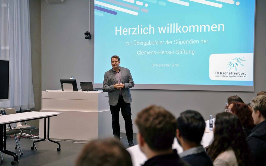 Mann im grauen Sacko steht vor blauer Leinwand mit dem Schriftzug Herzlich willkommen