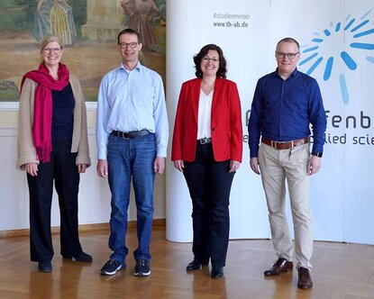 zwei Professorinnen und zwei Professoren stehen lächelnd nebeneinander. Im Hintergrund links ist der Teil eines Ölgemäldes zu sehen, rechts im Hintergrund ein Teil der weißen Messewand mit dem blauen TH-Logo.