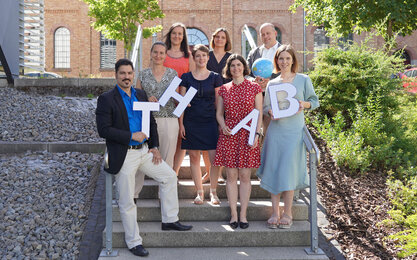 Gruppenfoto der Mitglieder des International Offices der TH AB, Personen stehen auf einer Steintreppe im Freien und halten Pappbuchstaben TH AB in der Hand.