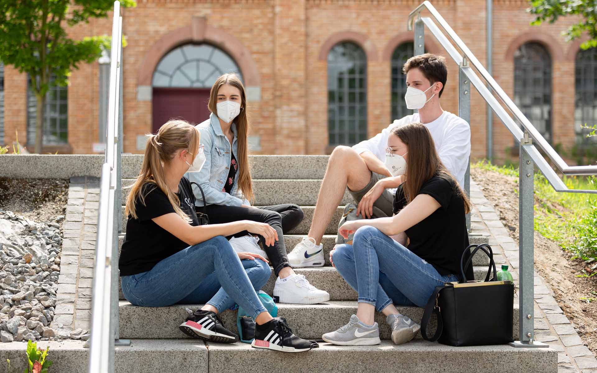 Studierende mit Maske sitzen auf einer Treppe am Campus 1 und unterhalten sich