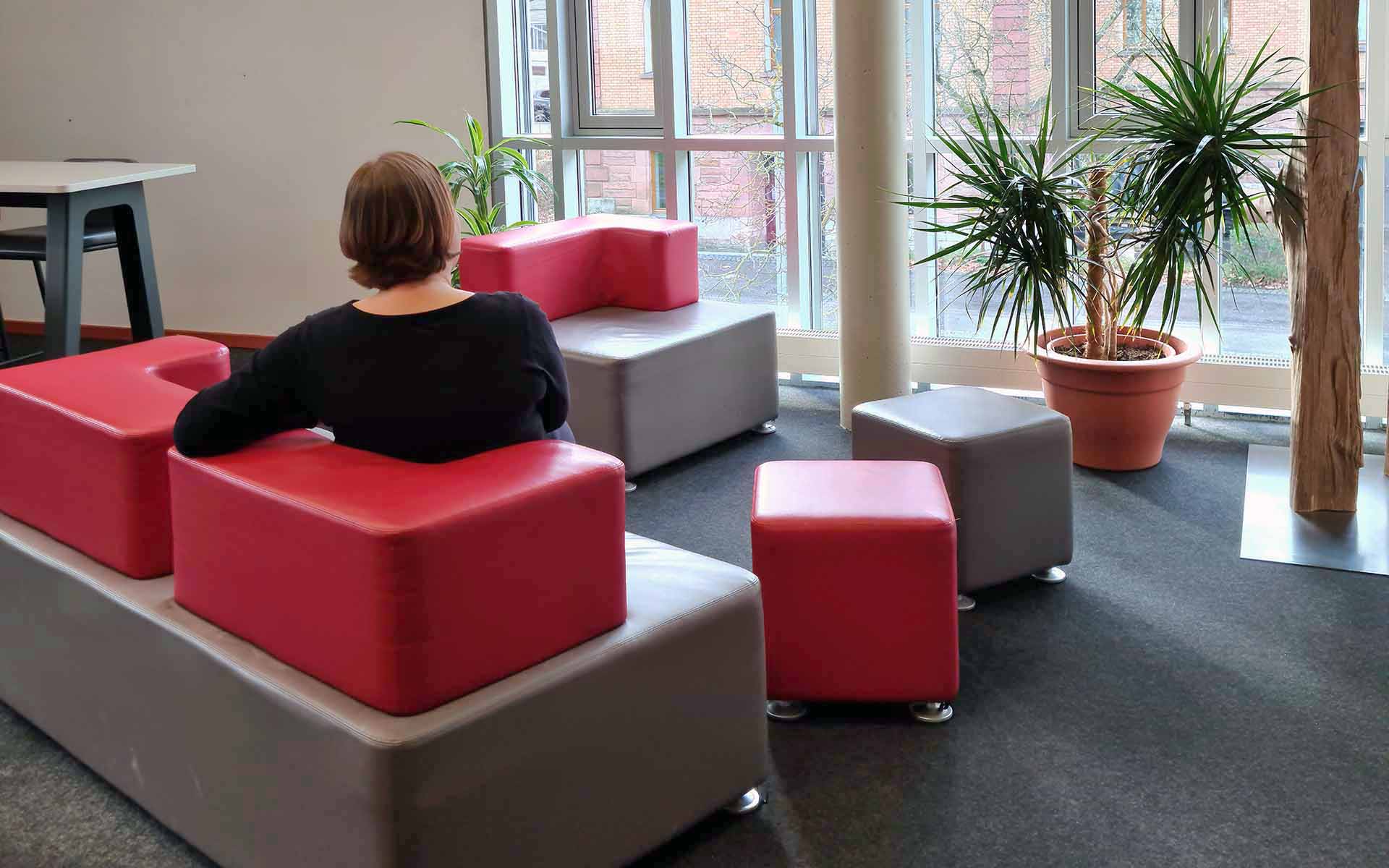 Ein rot-braunes Sofa, auf dem eine Frau in schwarzem Pullover sitzt und zum Fenster rausschaut, vor dem eine Topfpflanze steht