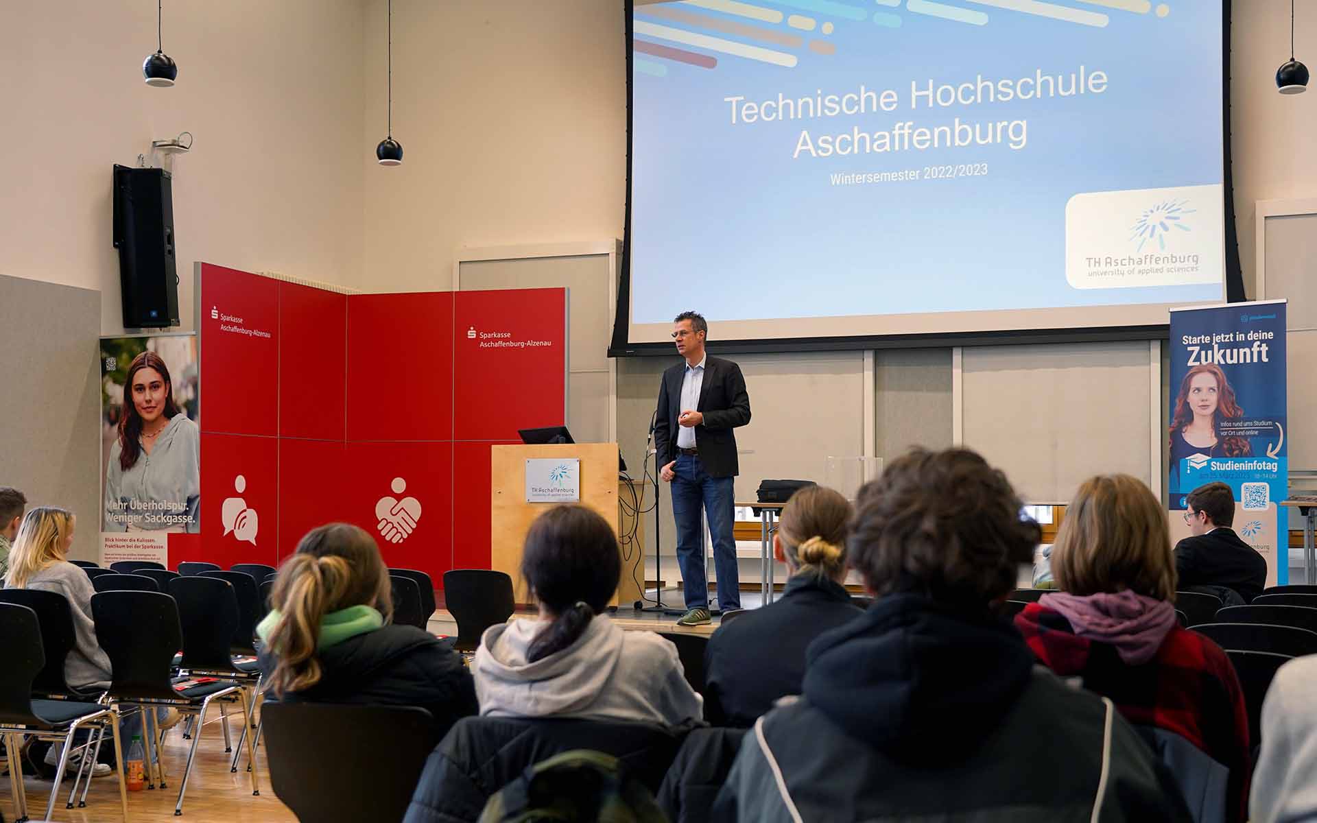 Ein Professor der Fakultät Wirtschaft und Recht steht in der Aula neben dem Rednerpult und spricht zu den sitzenden Schülerinnen und Schülern. Im Hintergrund ist auf einer blauen Leindwand der Schriftzug der TH Aschaffenburg zu sehen. Daneben steht einer rote Wand der Sparkasse.