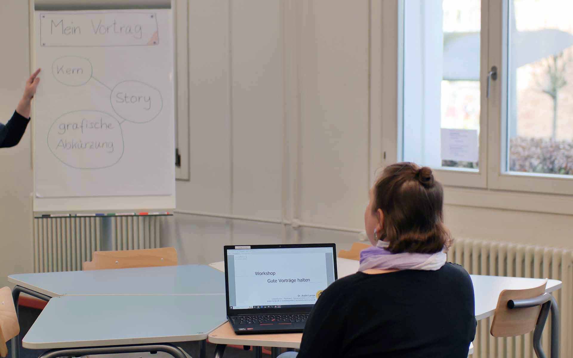 Eine Frau sitzt vor einem aufgeklappten Laptop mit Blick auf ein Flipchart