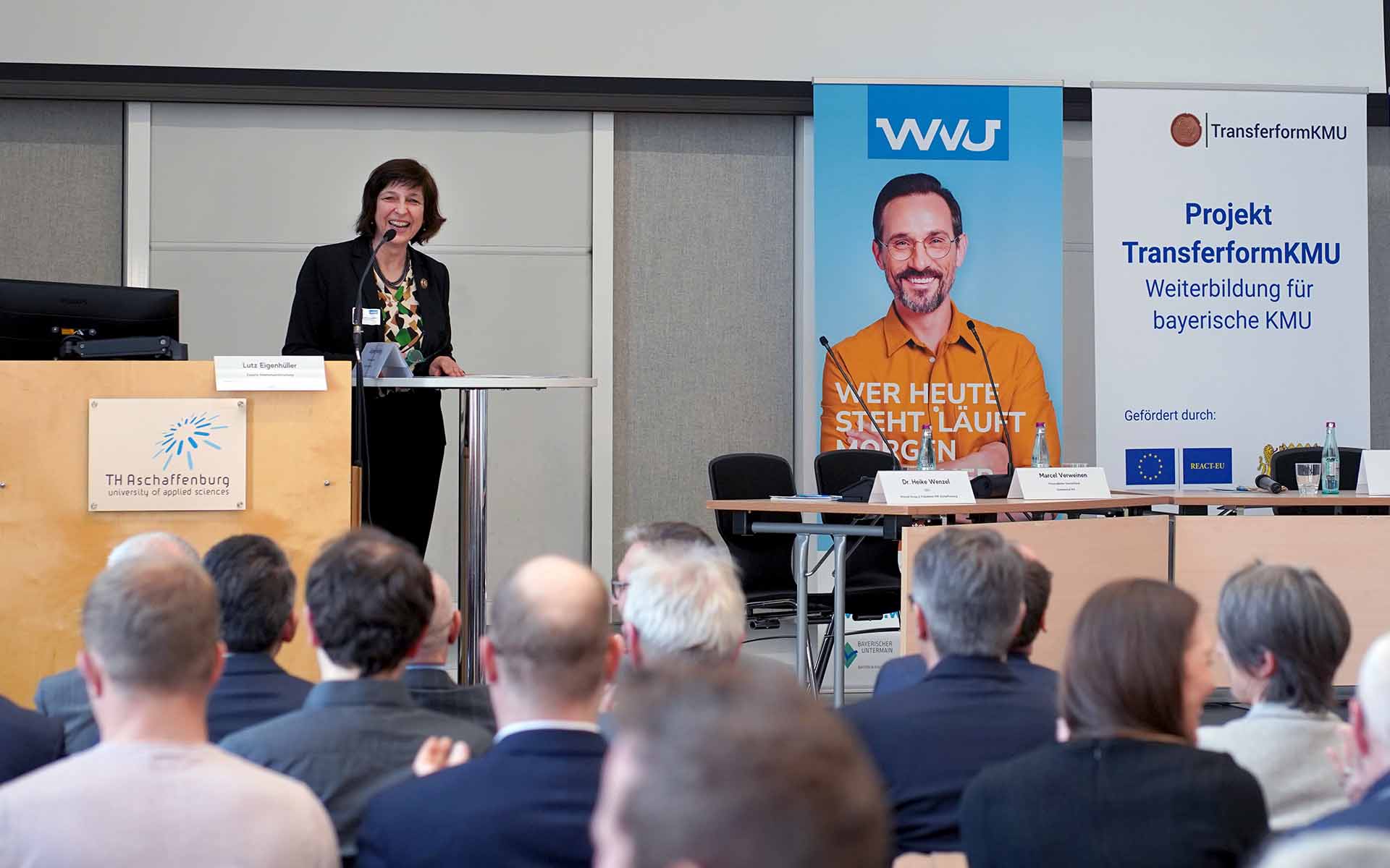 Die TH-Präsidentin steht lächelnd am Rednerpult mit Mikrophon. Das sitzende Publikum ist von hinten zu sehen. Rechts hängt ein Plakat des WVU mit dem Porträt eines jungen Mannes in orangefarbenen Hemd.