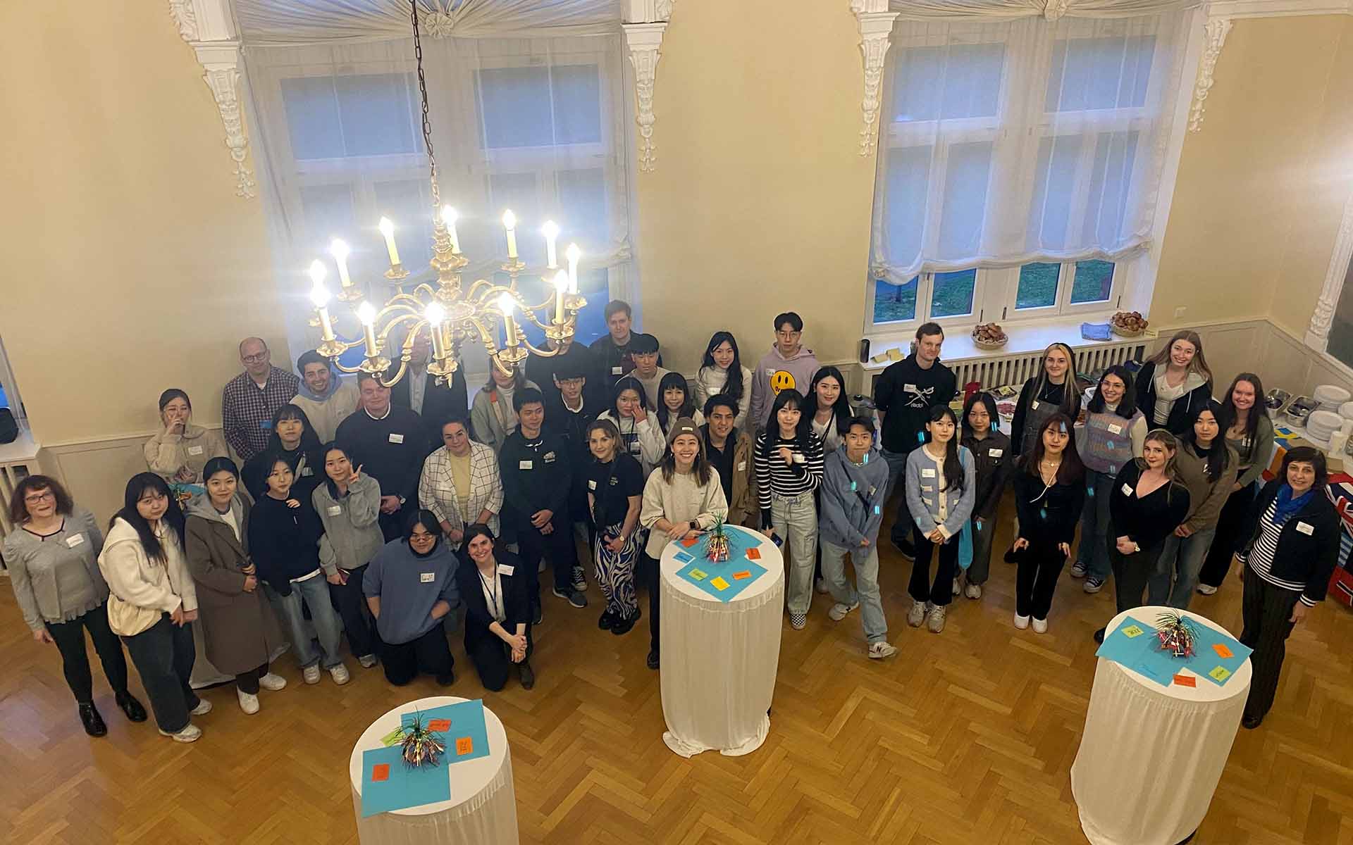 Die Gruppe der Austauschstudierenden steht vor den Fenstern des Hocksaals, der mit einem Kronleuchter beleuchtet ist, mit Blick zur Kamera hinter drei Stehtischen