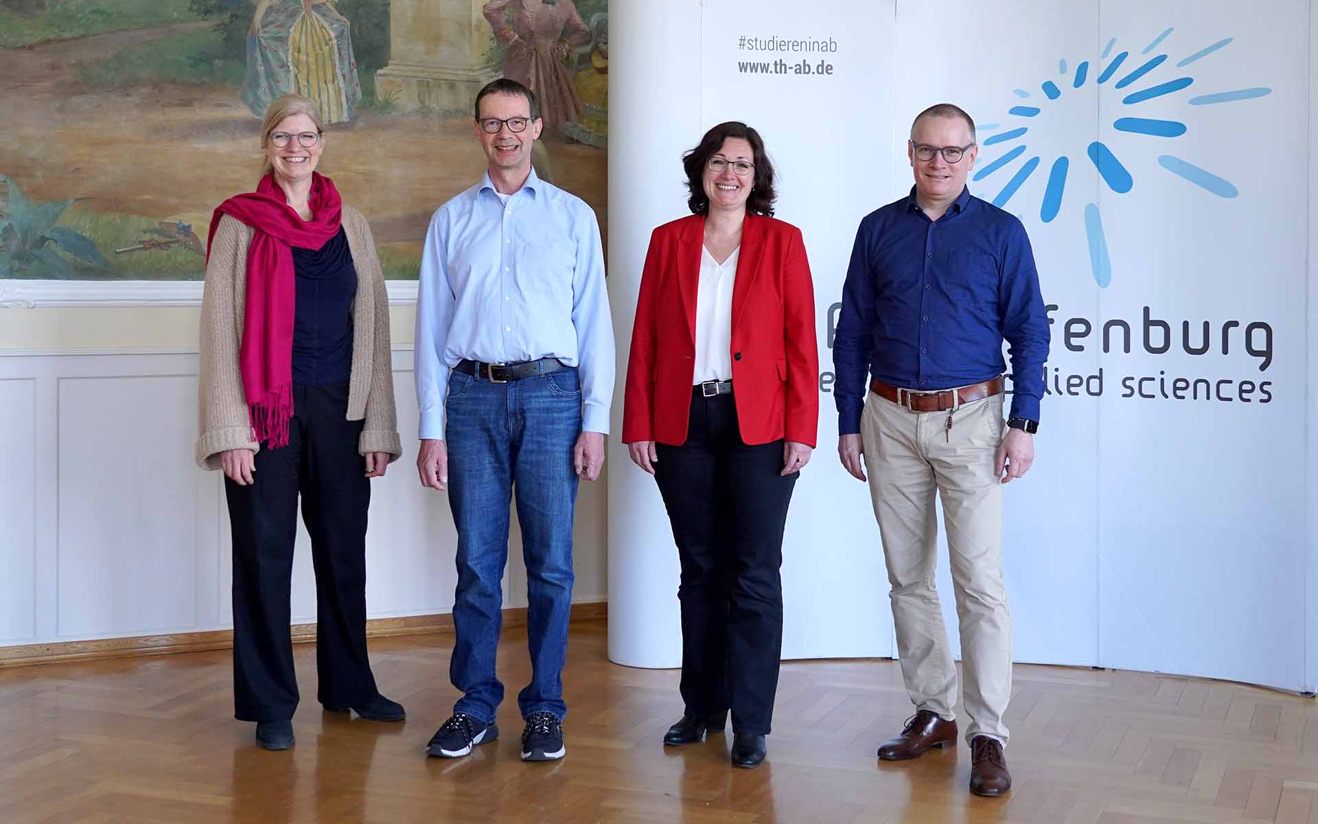 zwei Professorinnen und zwei Professoren stehen lächelnd nebeneinander. Im Hintergrund links ist der Teil eines Ölgemäldes zu sehen, rechts im Hintergrund ein Teil der weißen Messewand mit dem blauen TH-Logo.