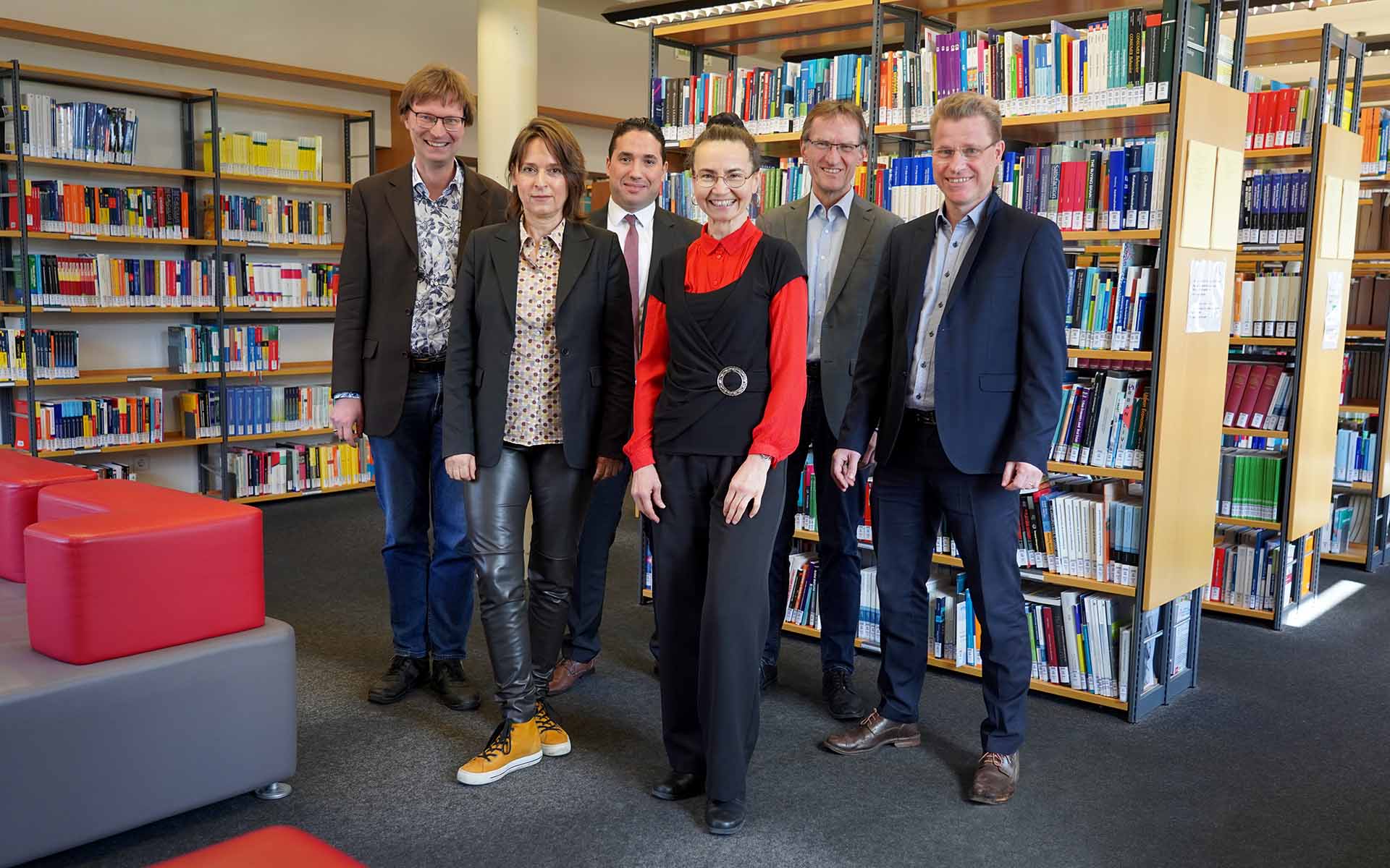 Zwei Professorinnen und vier Professoren stehen nebeneinander vor Bücherregalen in der Bibliothek und lächeln in die Kamera