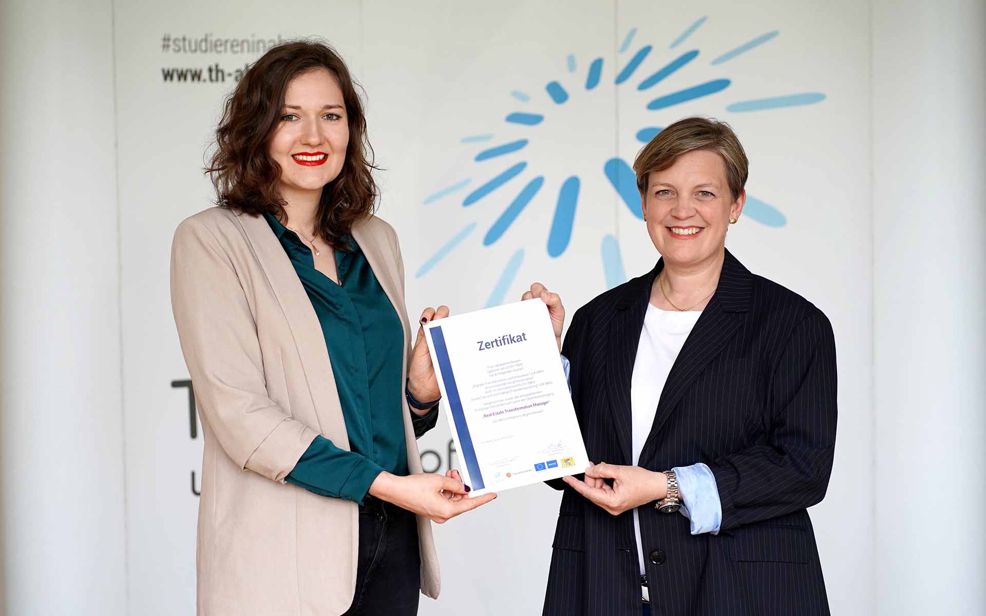 Die Absolventin hält ihr Zertifikat in den Händen, das sie von der Professorin überreicht bekommt. Auf der weißen Wand im Hintergrund ist das blaue TH-Logo zu sehen.