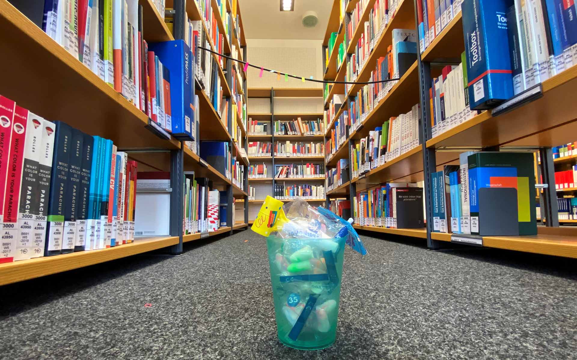 auf dem Boden vor mehreren Bücherregalen steht ein türkisfarbener Becher, der mit Ohrenstöpseln und einem Papierschirmchen gefüllt ist