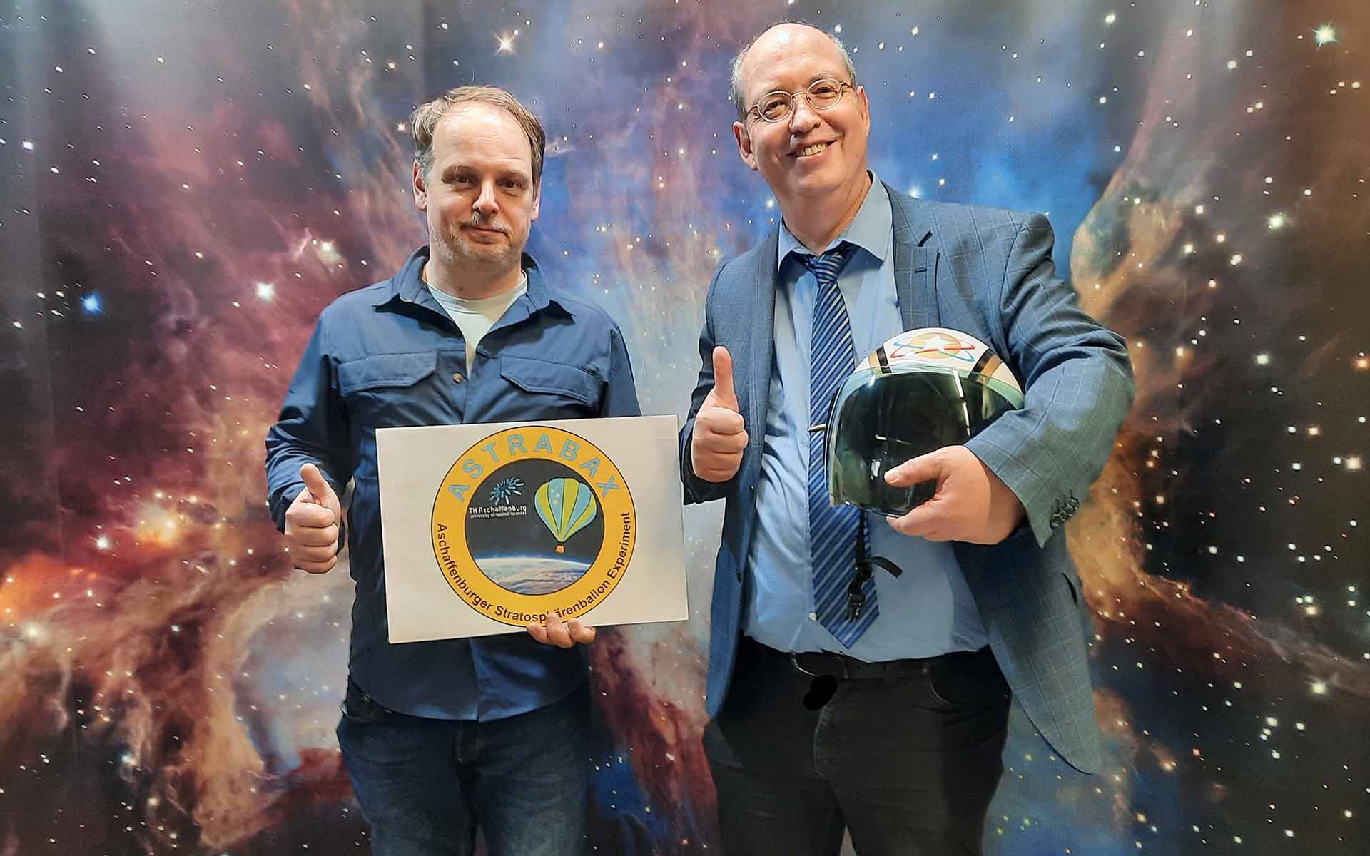 Professor Hildenbrand hält das Logo des Projekts in der Hand, Professor Döhring einen Astronautenhelm. Beide heben den Daumen der rechten Hand in die Höhe und stehen vor einer Wand, die einen Sternenhimmel zeigt