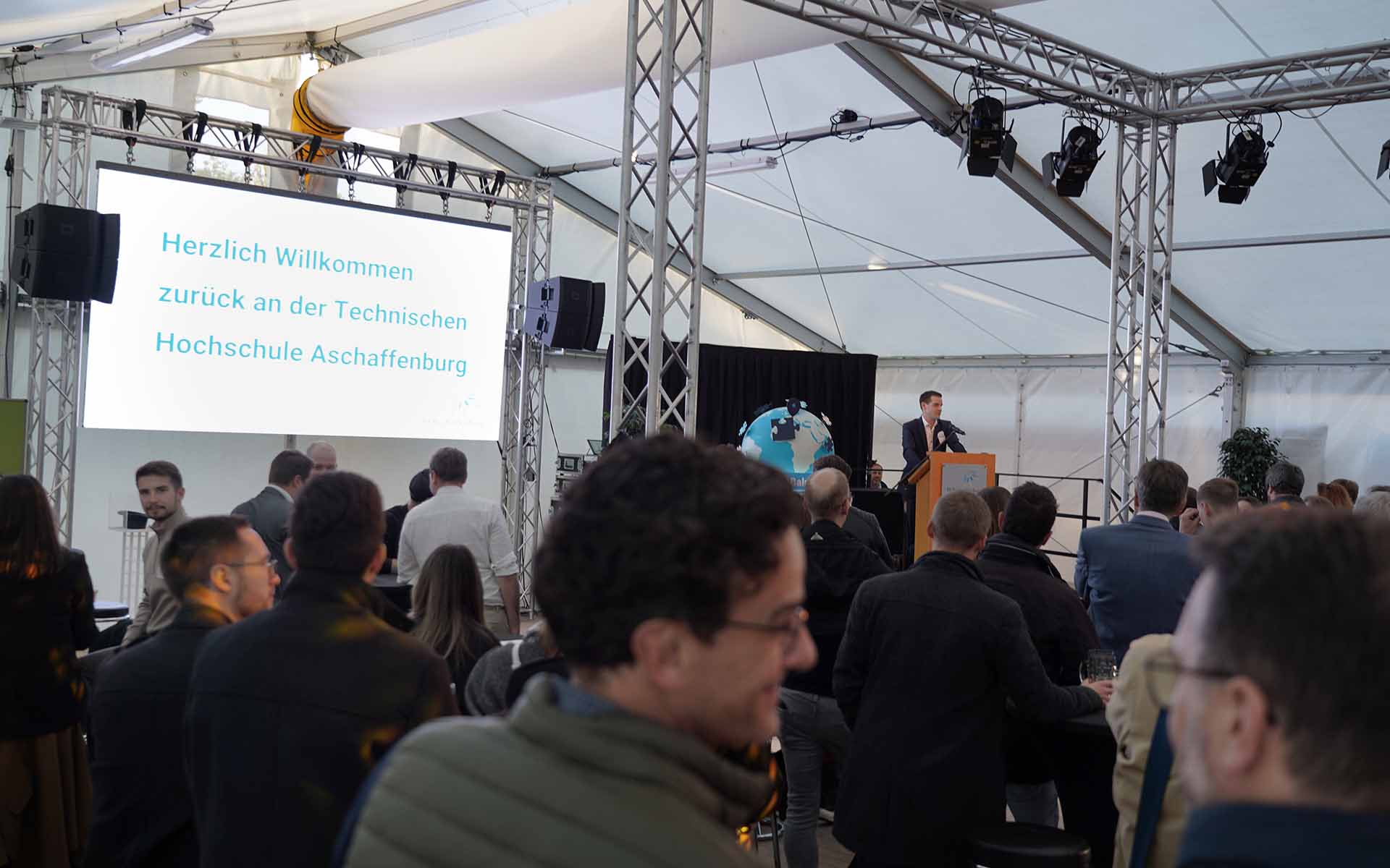 Mehrere Alumni von hinten oder der Seite im Zelt hören dem Vortrag des Alumni-Vorsitzenden am Rednerpult zu, Leinwand mit blauer Aufschrift "Herzlich willkommen zurück an der Technischen Hochschule Aschaffenburg" links im Hintergrund