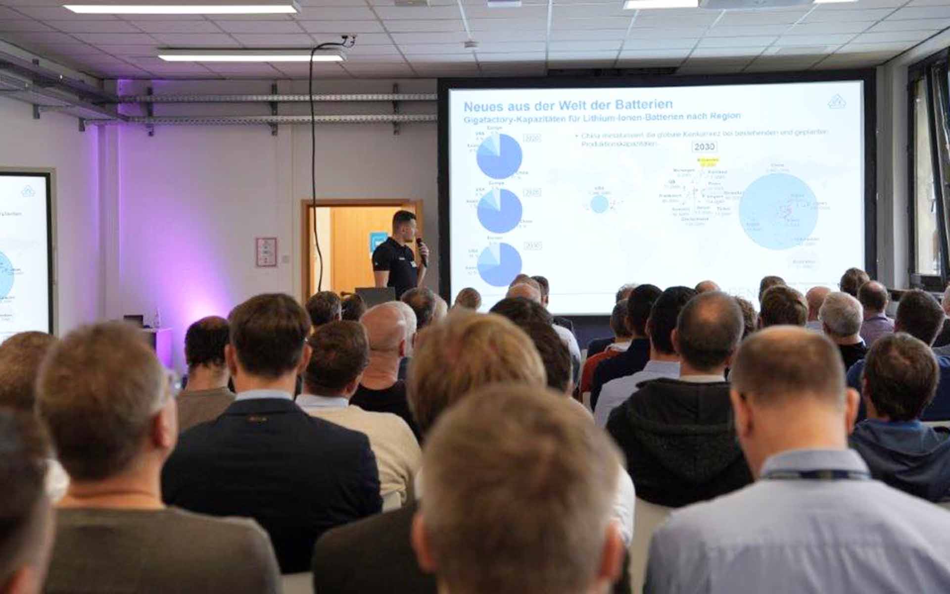 In Reihen sitzende Menschen von hinten mit Blick auf einen Referenten und eine Präsentation.