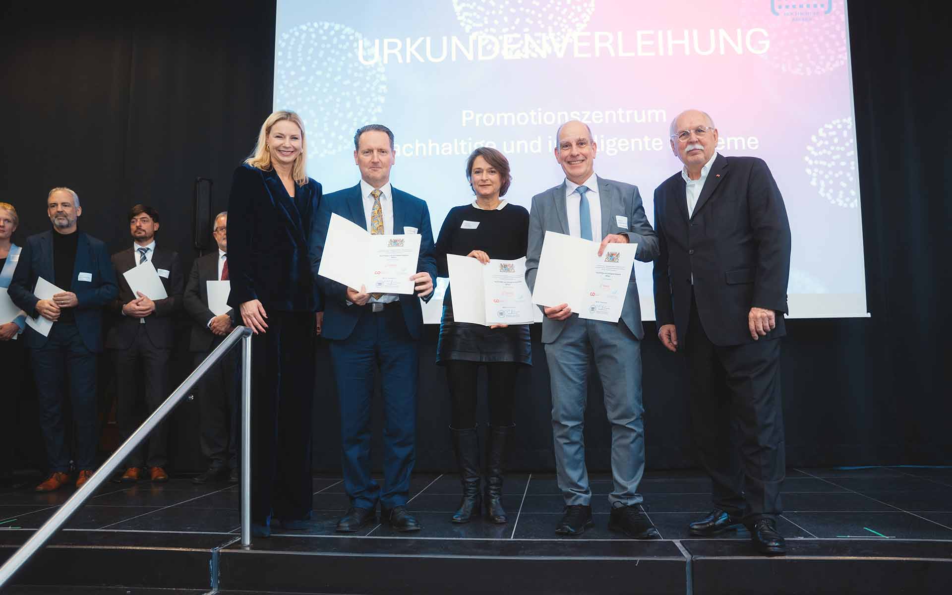 Fünf Menschen stehen vor einer Leinwand mit dem Schriftzug Urkundenverleihung. Drei Personen halten Urkunden in den Händen.
