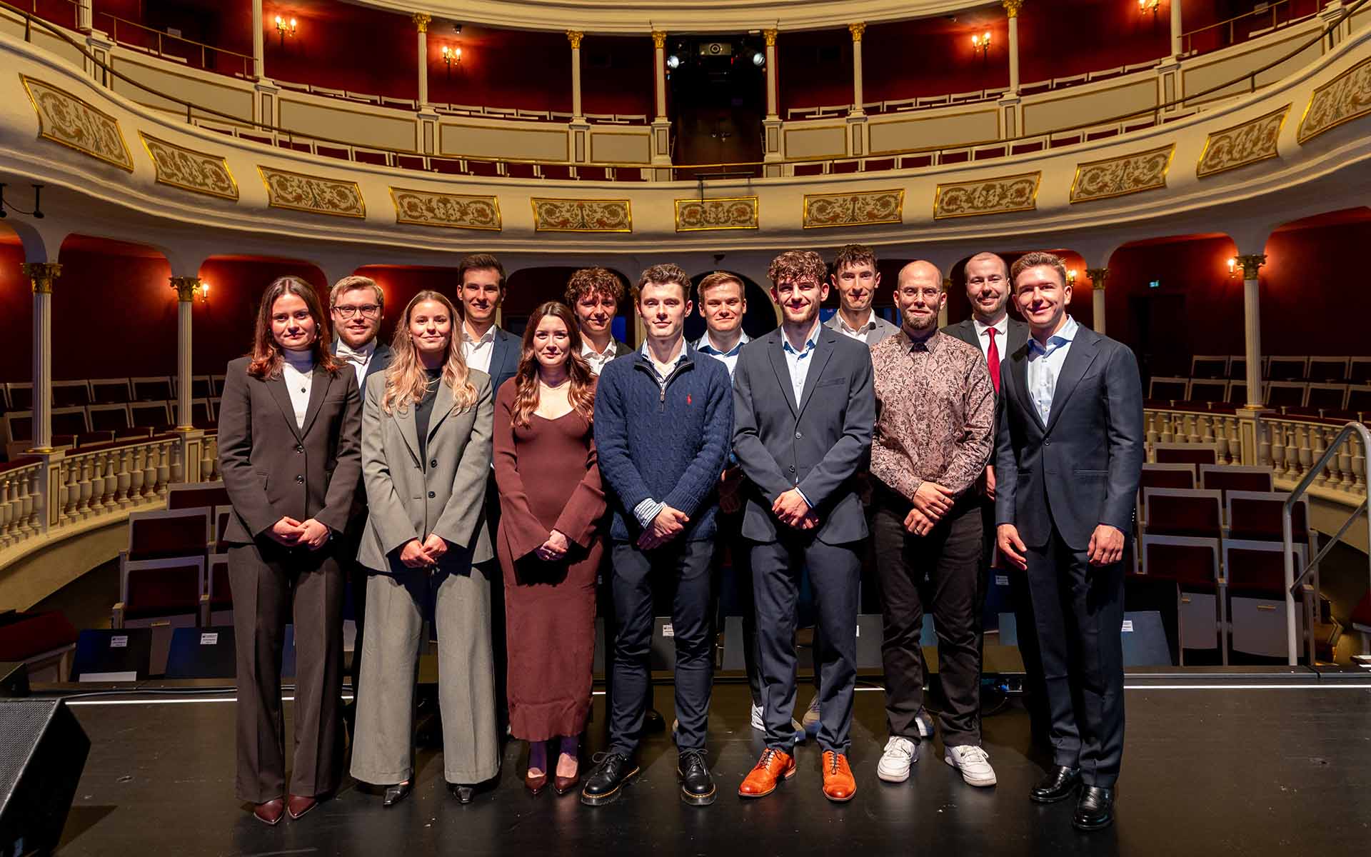 Gruppenbild mit jungen Männern und Frauen in Abendgarderobe auf der Bühne vor Zuschauerrängen im Theater