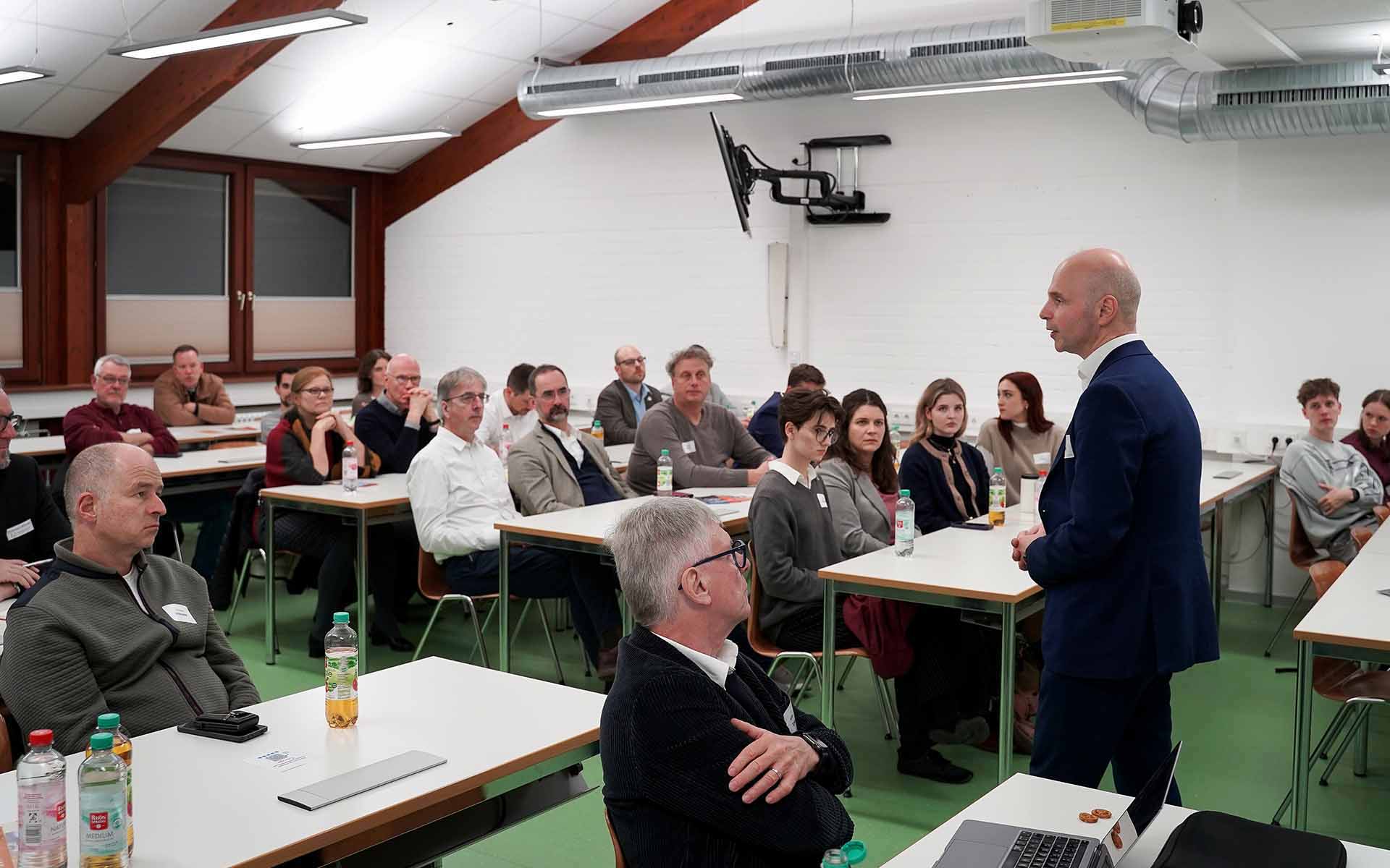 Mann im schwarzen Anzug steht im Hörsaal vor Teilnehmenden an Tischen und hält einen Vortrag