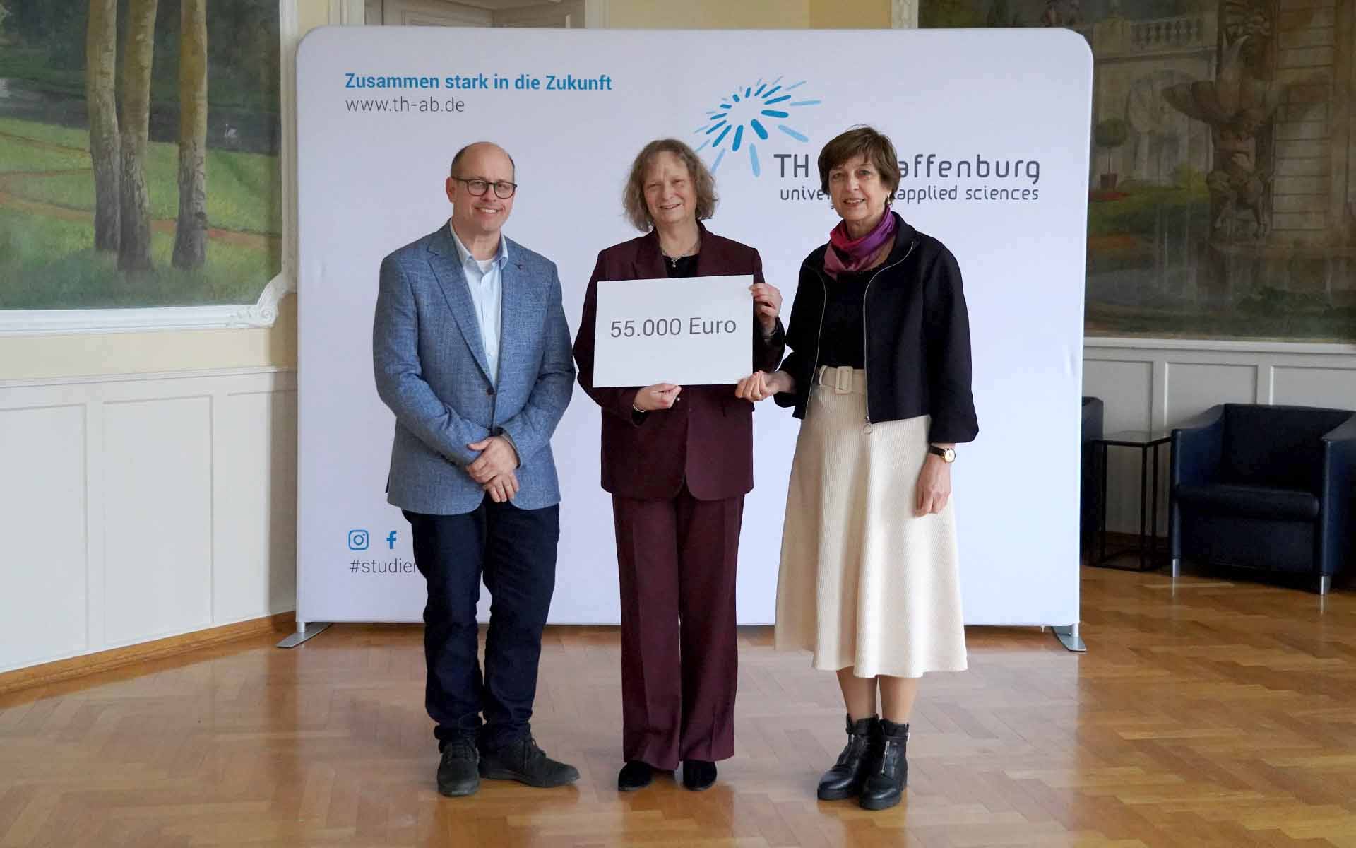 Frau in der Mitte hält Schild mit Spendensumme, Mann und Frau stehen rechts und links neben ihr vor TH-Logowand