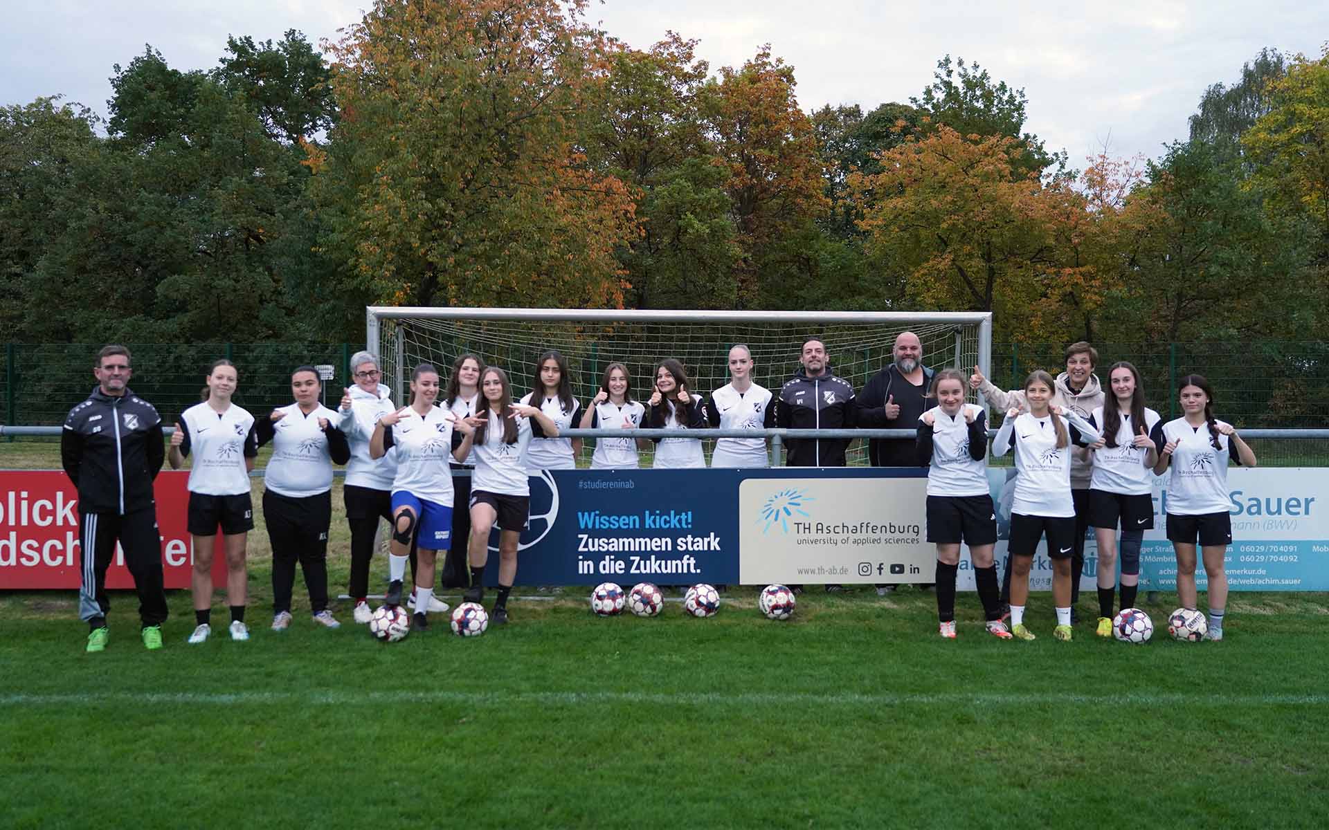 Fußballmannschaf mit Trainern steht vor Bandenwerbung und Tor auf dem Fußballplatz