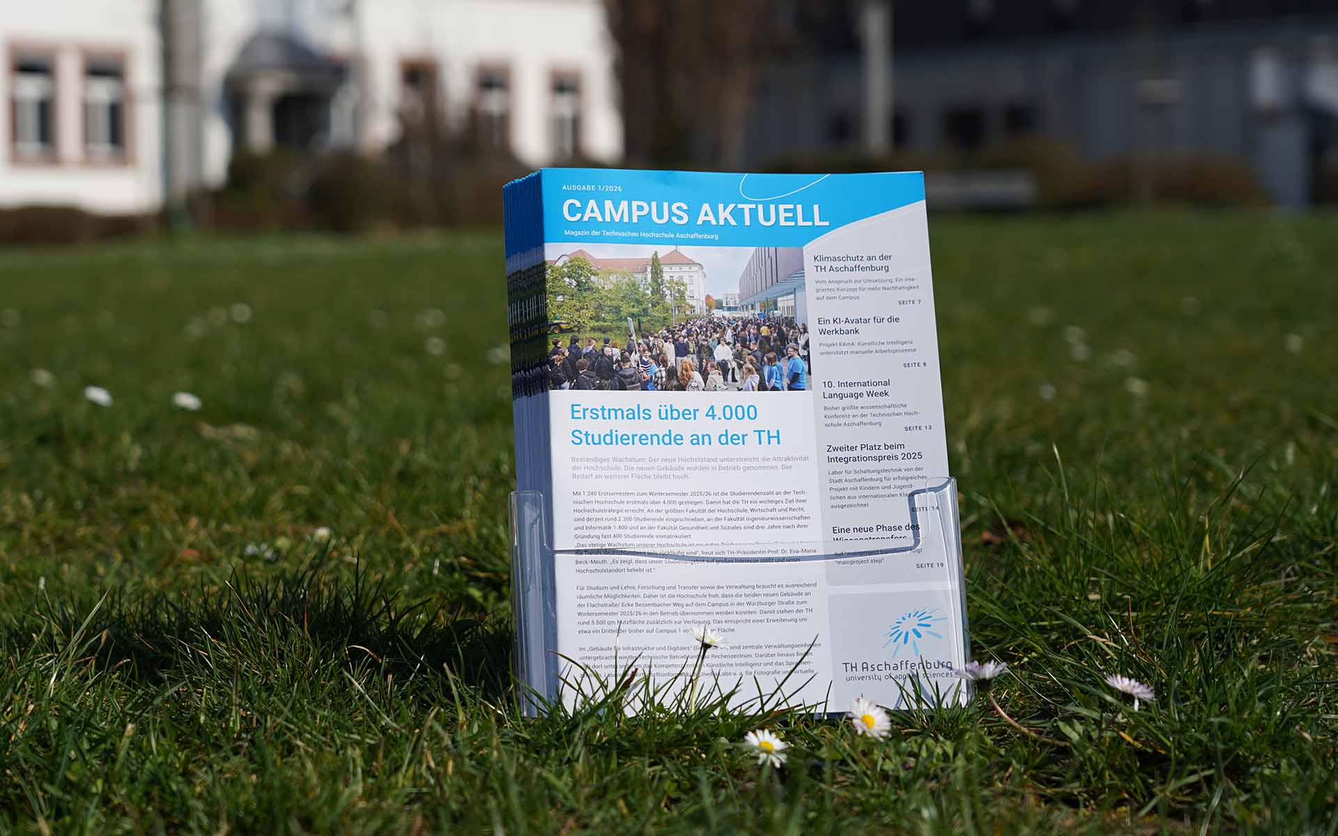 ochschulmagazine im Prospektständer auf Wiese mit Gänseblümchen vor Gebäuden der TH