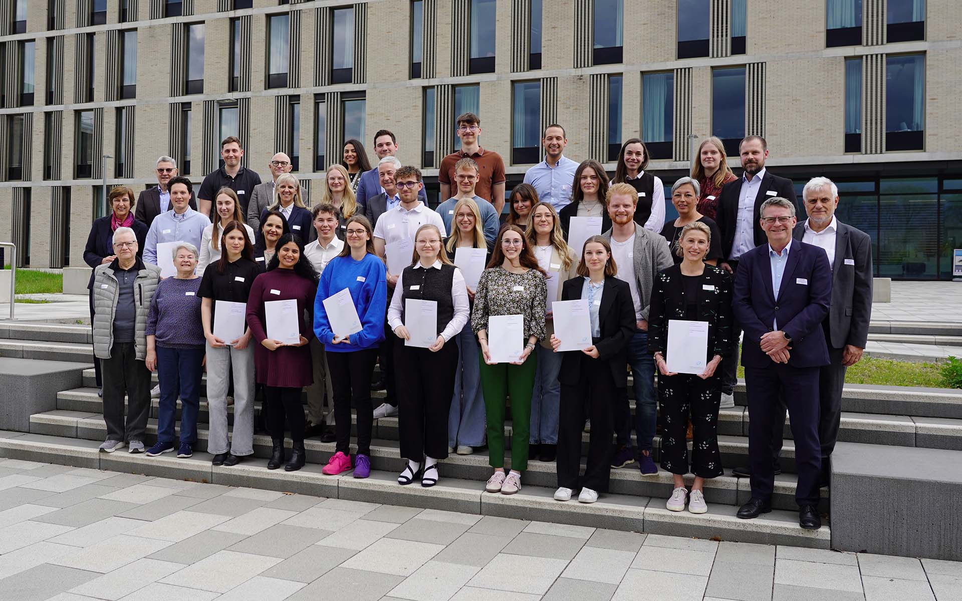 Gruppenbild mit rund 40 Personen, die teilweise Zertifikate halten, auf Treppenstufen vor den Neubauten