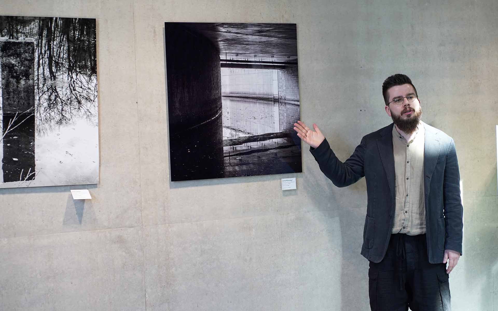 Der Künstler steht vor einer Fotografie an der Wand und zeigt mit der Hand darauf.