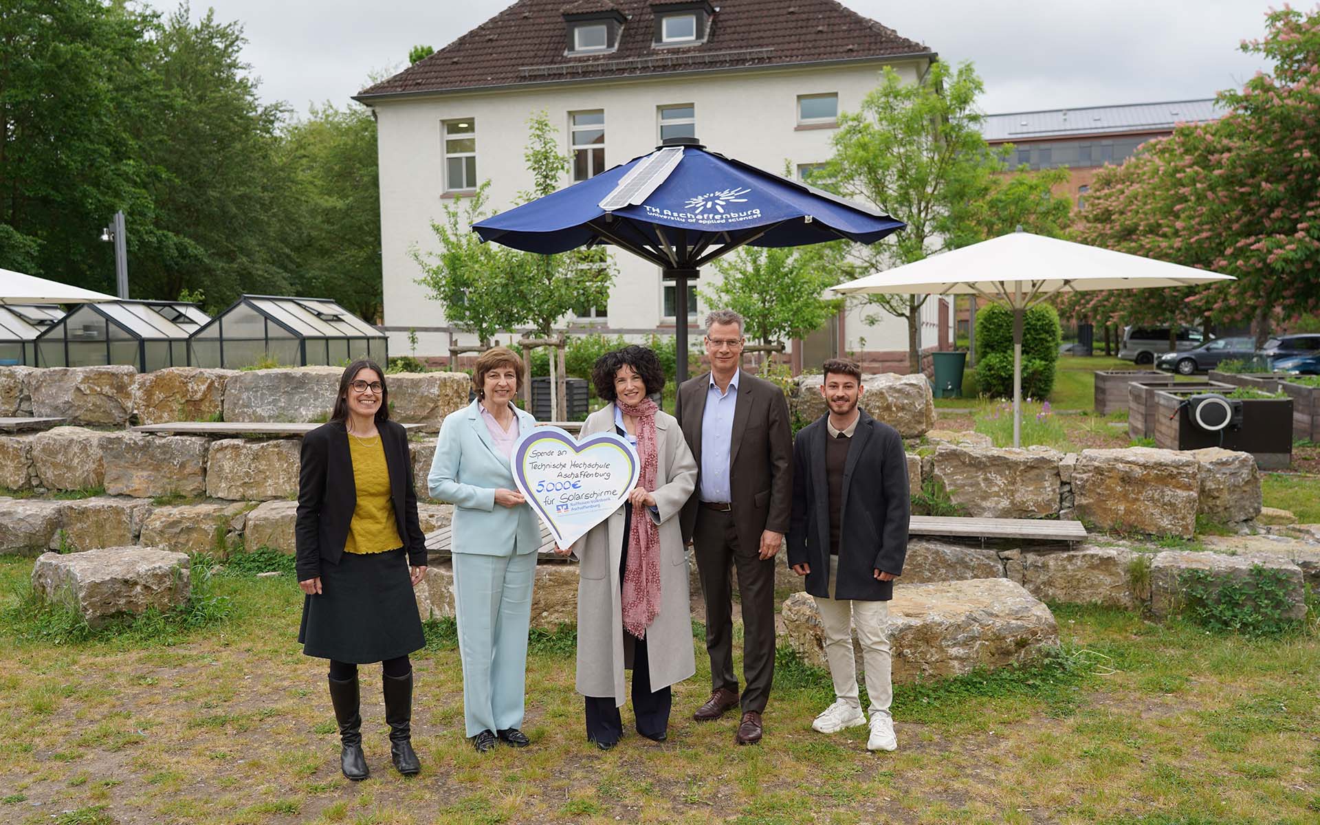 Frau mit herzförmigem Schild mit Spendensumme steht zwischen zwei Frauen und zwei Männern vor geöffnetem blauen Solarschirm