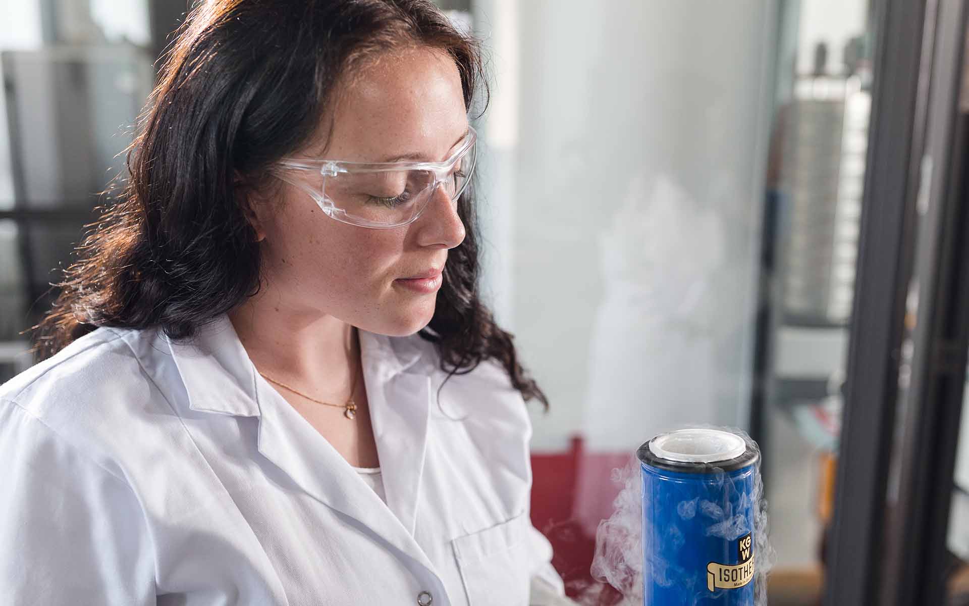 Studentin mit Schutzbrille und weißem Kittel hält ein Gefäß mit Stickstoff in der Hand