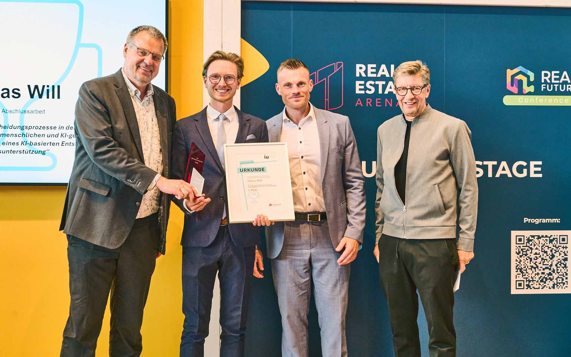 Gewinner mit Urkunde und drei weiteren Männern auf der Bühne