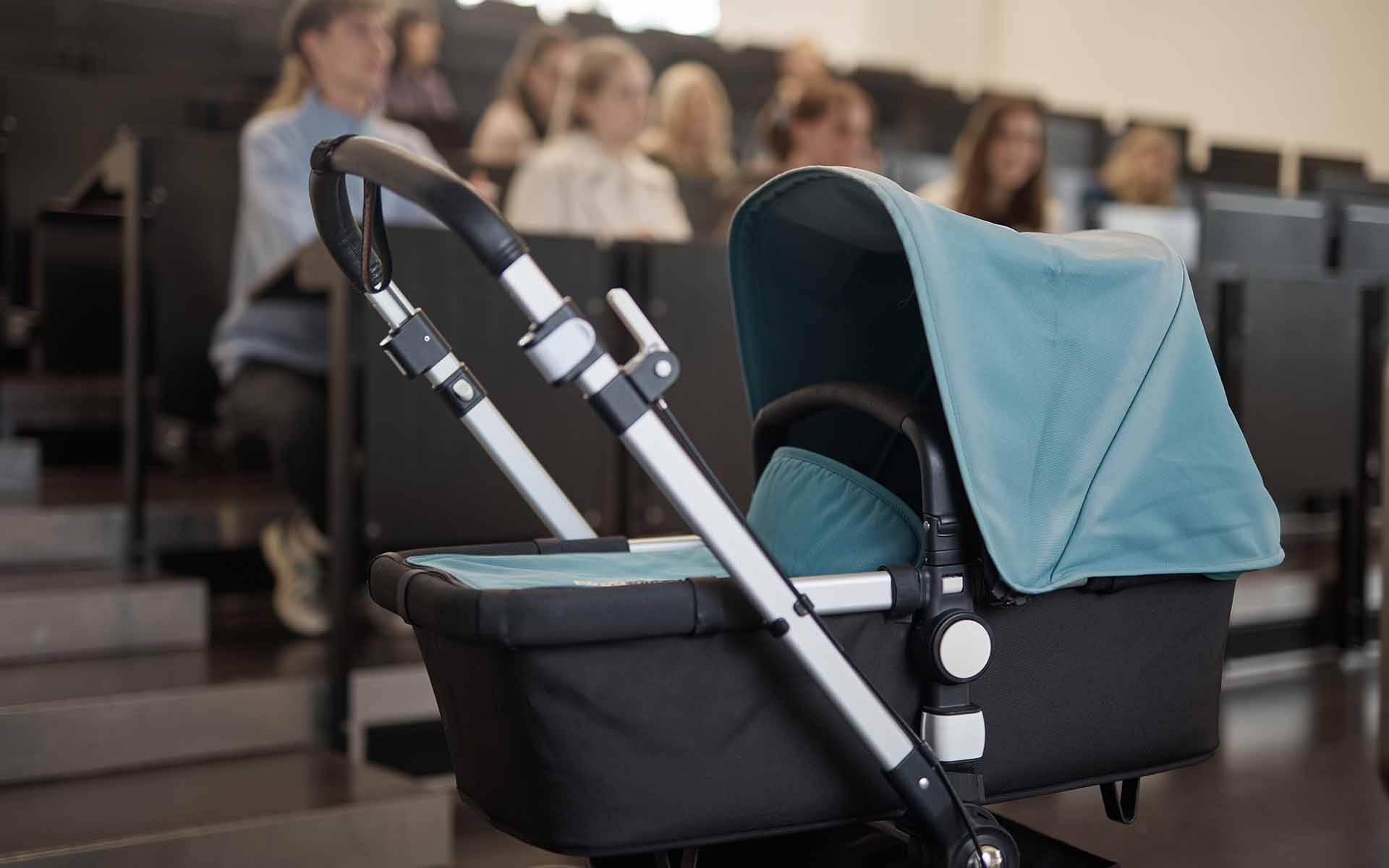 Kinderwagen steht im Hörsaal vor ansteigenden Reihen mit Studierenden