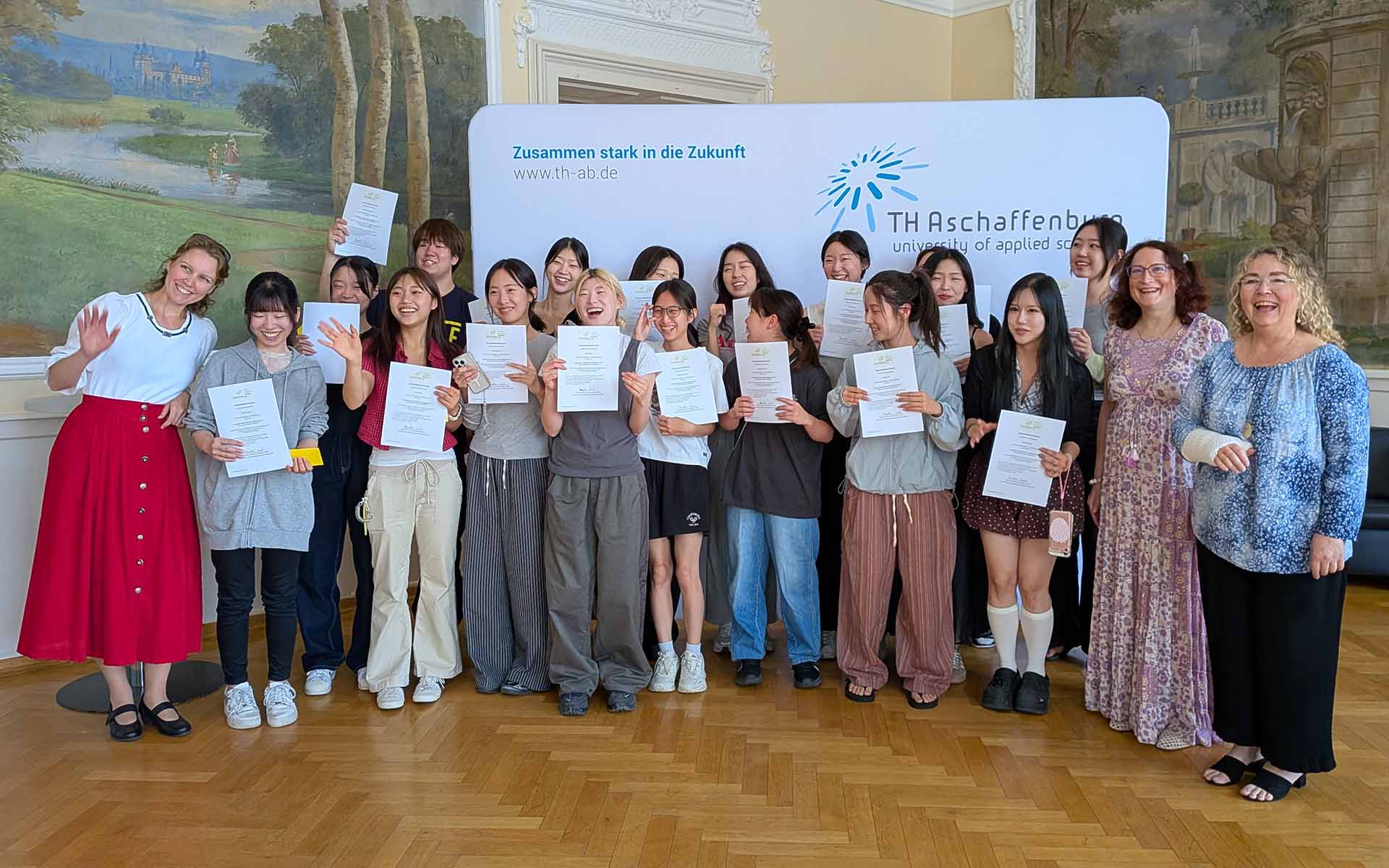 Gruppenfoto mit 20 Personen mit Zertifikaten im Hocksaal vor Logowand