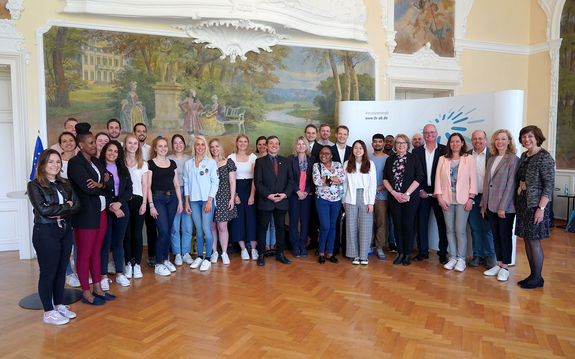 Eine Gruppe Studierenden sowie Professorinnen und Professoren in einem großen Saal der Hochschule vor einem Wandgemälde 