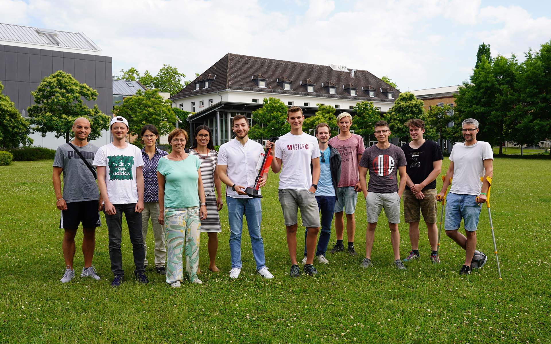 Gruppe von zwölf Personen steht auf einer grünen Wiese vor einem Gebäude und grünen Bäumen