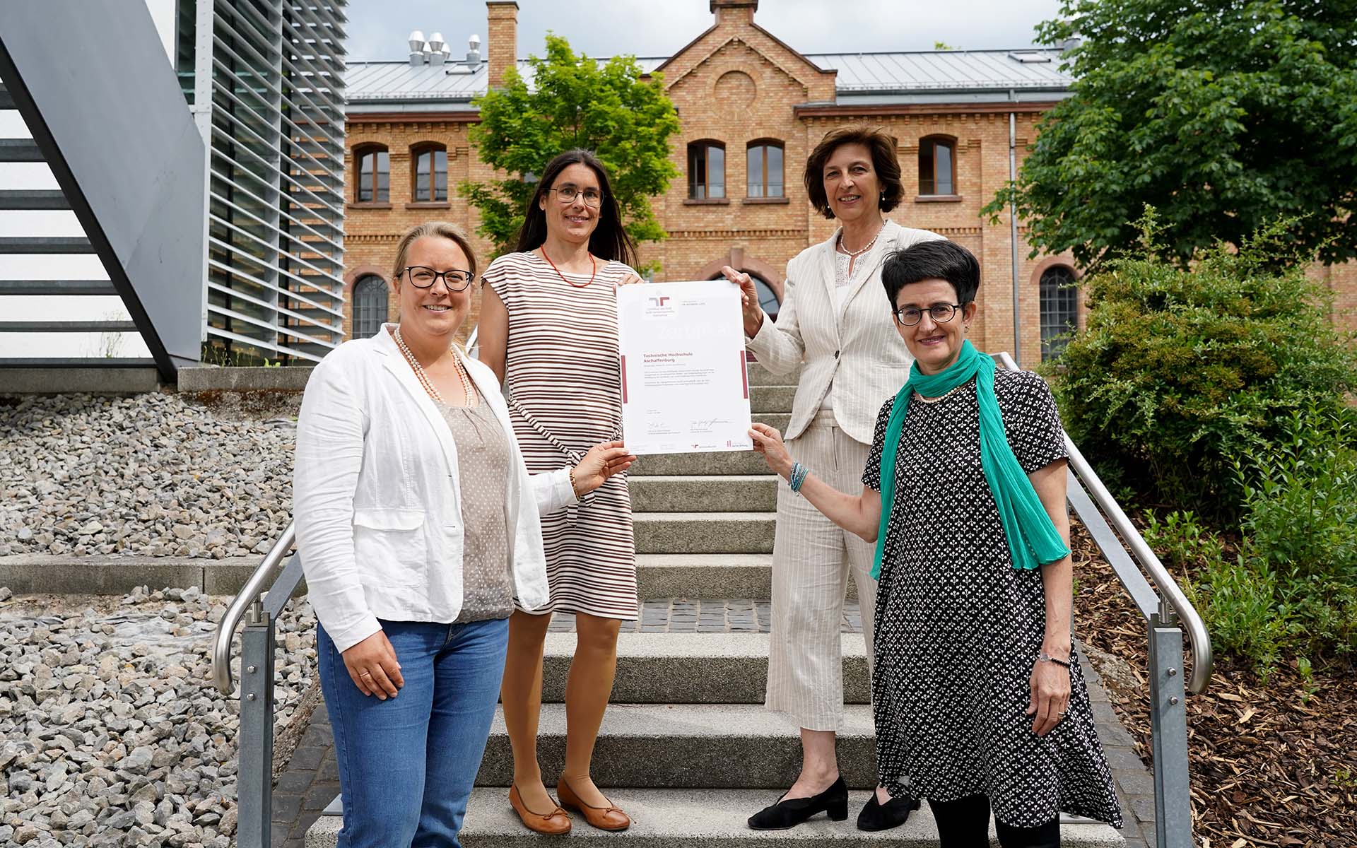 Vier Frauen stehen lächelnd auf einer Treppe vor einem Gebäude und halten ein Blatt Papier fest