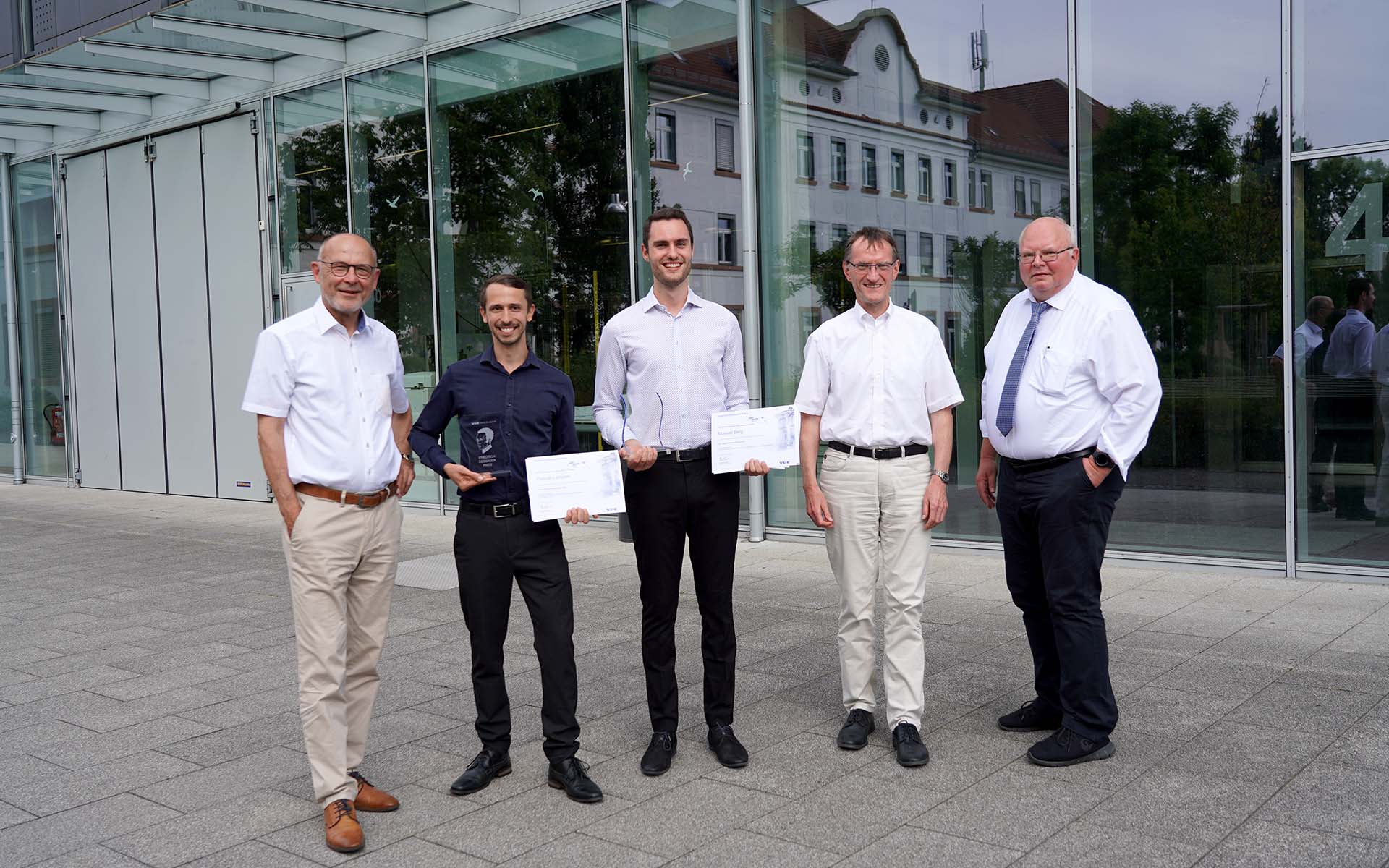 Fünf Männer - darunter die beiden Preisträger und die betreuenden Professoren - stehen lächelnd nebeneinander vor einem Gebäude der Hochschule mit spiegelnder Glasfassade. Die beiden Preisträger halten ihre Urkunden und den Preis in den Händen.