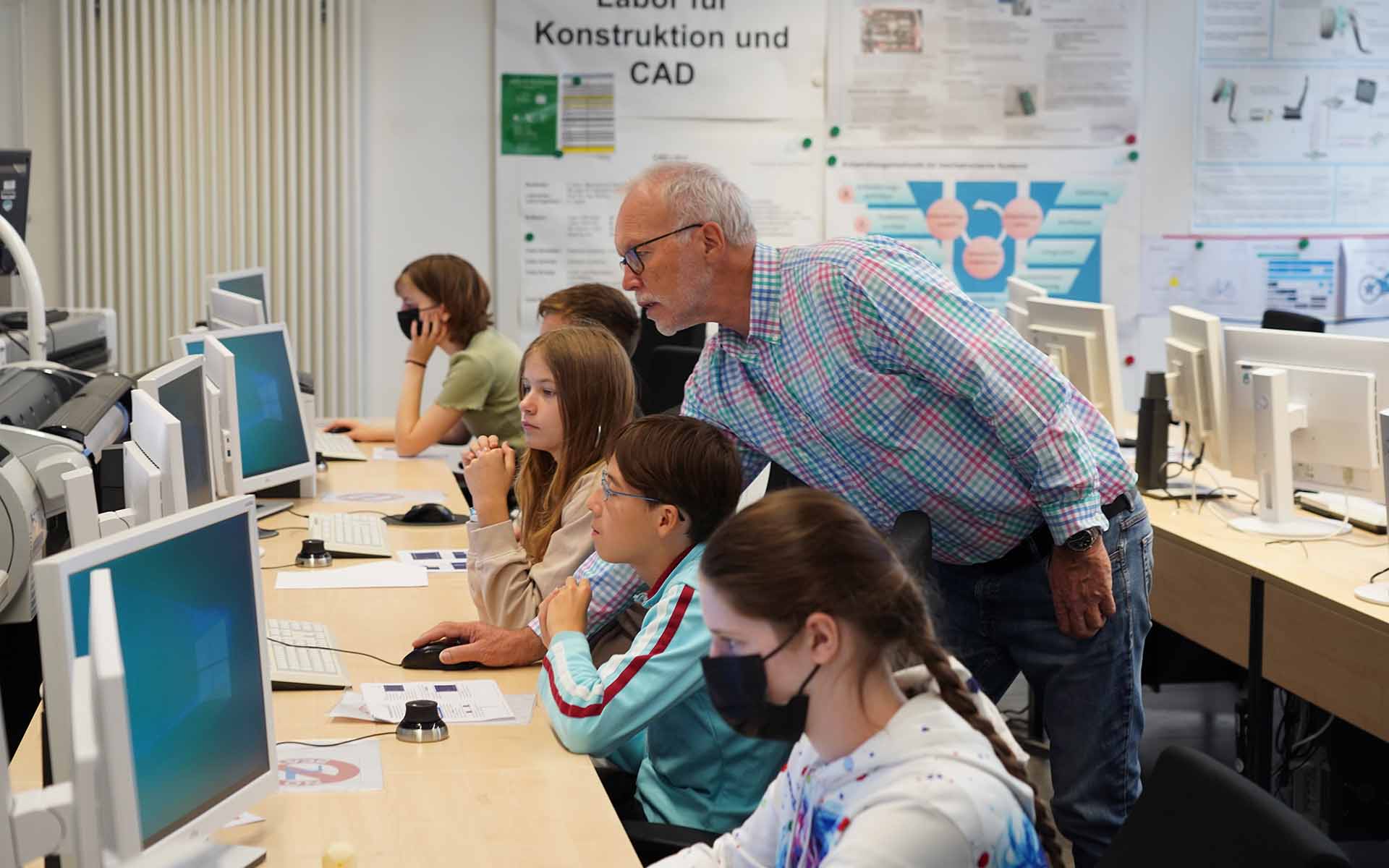 Ein Mann steht hinter fünf Schülerinnen und Schülern, die vor Bildschirmen sitzen und zeigt ihnen etwas mit der Computermaus