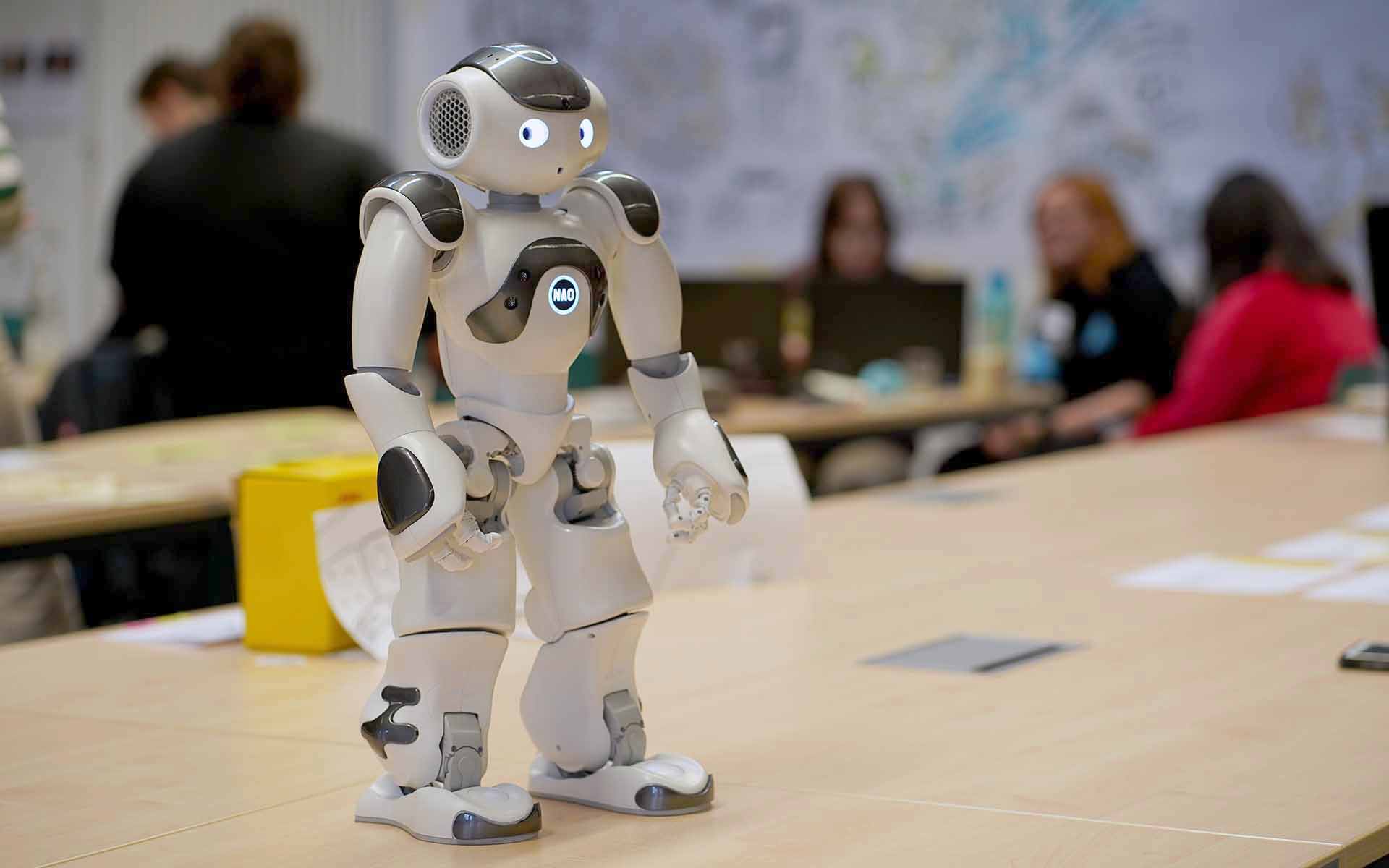 Menschenähnlicher Roboter steht auf einem Tisch im Hörsaal und im Hintergrund sind einige Schülerinnen bei der Gruppenarbeit zu sehen