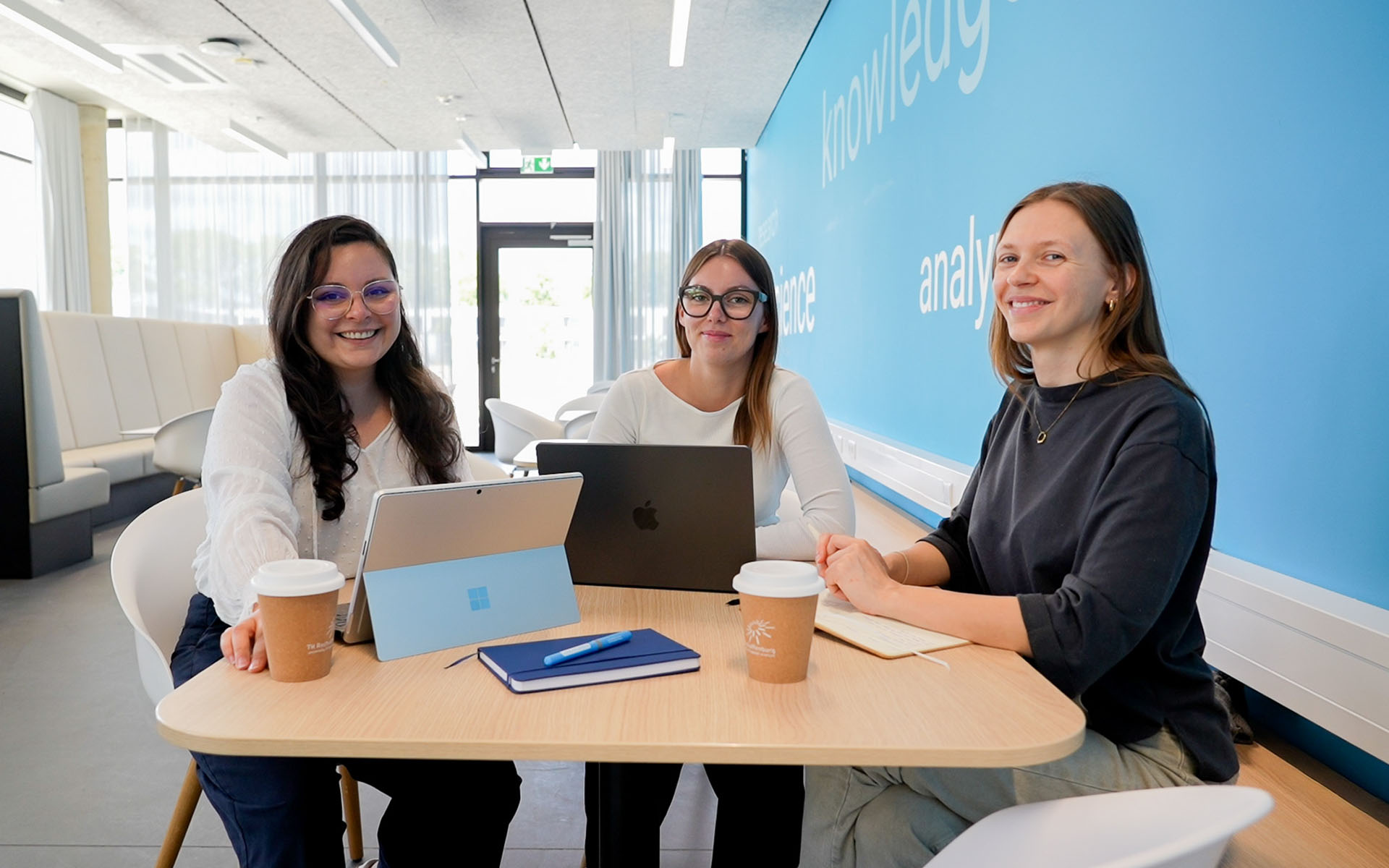 Drei Studierende sitzen an einem kleinen Tisch in einem modernen Aufenthaltsraum und arbeiten gemeinsam an Laptops.